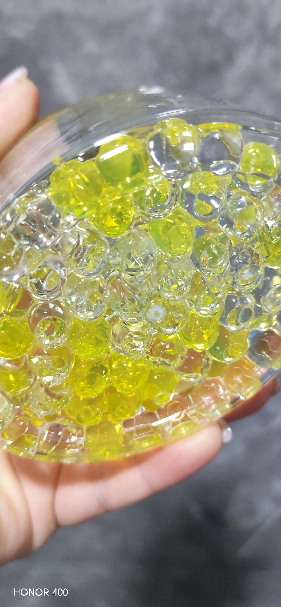 A close-up shot depicts a clear glass bowl held in a person's hand, filled with numerous translucent spheres, some yellow and some clear. The background is a blurry grey. The image includes a watermark "HONOR 400" in the bottom left corner. The scene does not appear to be located in any specific location and there is no indication this image was taken in the city of Noviy, Russia.