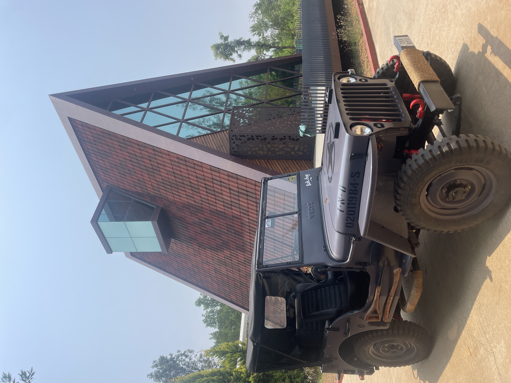 A grey, vintage-style Jeep, possibly a customized Willys, is parked on a paved surface next to a modern building under a clear, bright sky. The Jeep features a classic slotted grille, round headlights, and a front bumper equipped with red tow hooks and a rope-wrapped object, possibly part of a winch. "WILLYS" is stenciled on the hood, and a "Bad Boy" sticker is visible on the windshield frame. A vertical registration number "02011984-S" is painted on the side of the engine compartment, and a license plate "CVN 0957" is on the front bumper. The vehicle has rugged off-road tires, a black soft top that appears to be rolled back, and black seats. Tools resembling a shovel and an axe are strapped to the side near the rear wheel.
The building behind the Jeep showcases contemporary architecture with a prominent triangular or pyramidal roofline. Its lower facade is covered in reddish-brown tiles or patterned wooden shingles. The upper section features a large glass facade with a dark geometric grid, reflecting the sky and surrounding trees. A distinctive, small, cube-shaped glass protrusion extends from one side of the building. A dark metal screen with geometric cutouts runs along a portion of the structure. The setting is outdoors, brightly lit by sunlight, casting strong shadows on the ground. Trees and green foliage are visible around the building. No specific activity is depicted; the scene is static.
