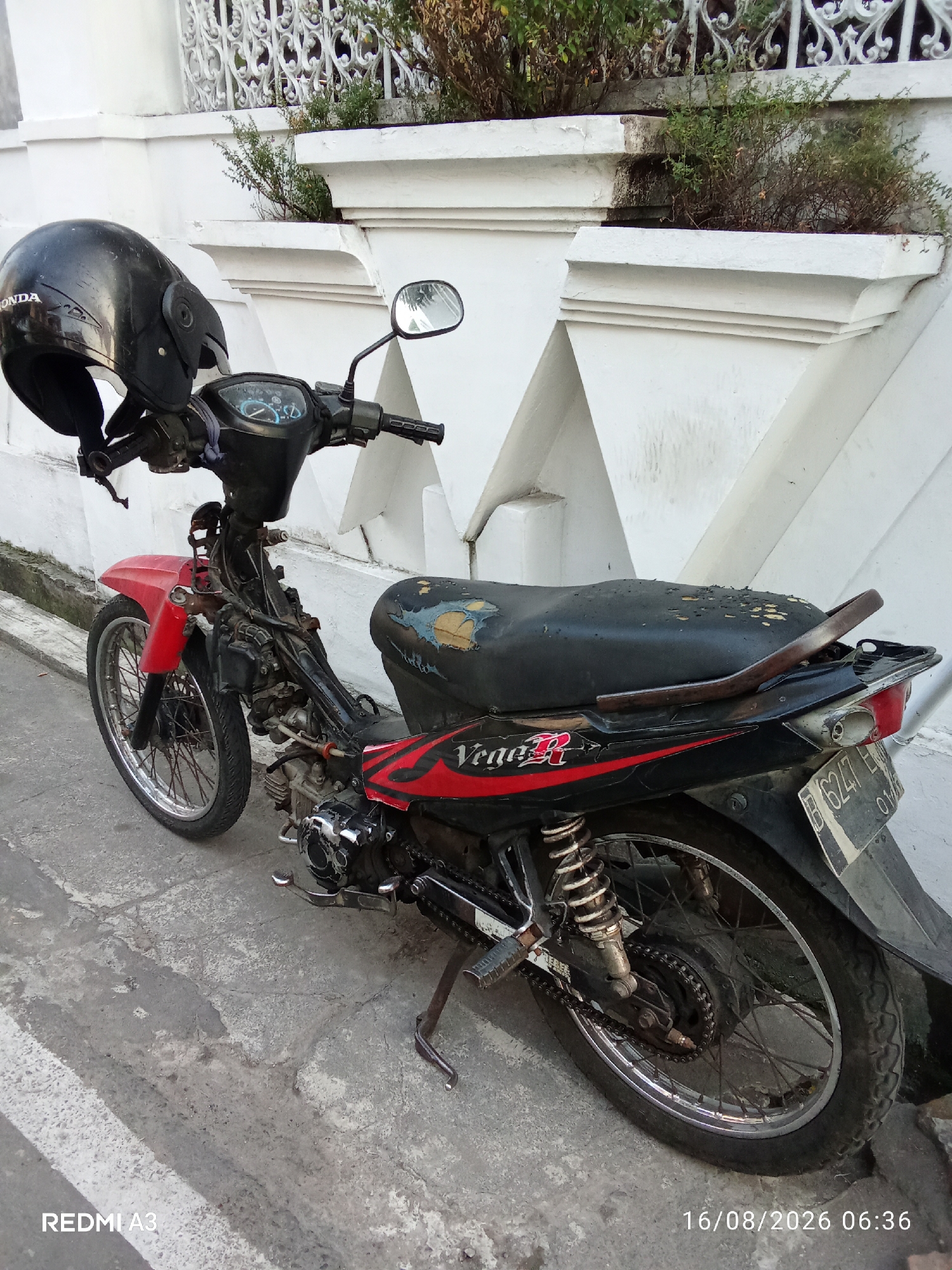 A parked motorcycle is in front of a white decorative wall with plants. The motorcycle is red and black, with a black helmet placed on the handlebars. The seat of the motorcycle is worn and torn, revealing the foam underneath. A license plate with the characters "B 6247 EH" is visible on the rear of the motorcycle. The ground is made of concrete. The image appears to be taken during the day, with lighting suggesting it is morning. The timestamp on the image indicates the date 16/08/2026 at 06:36. The location is identified as Banaran, Indonesia.