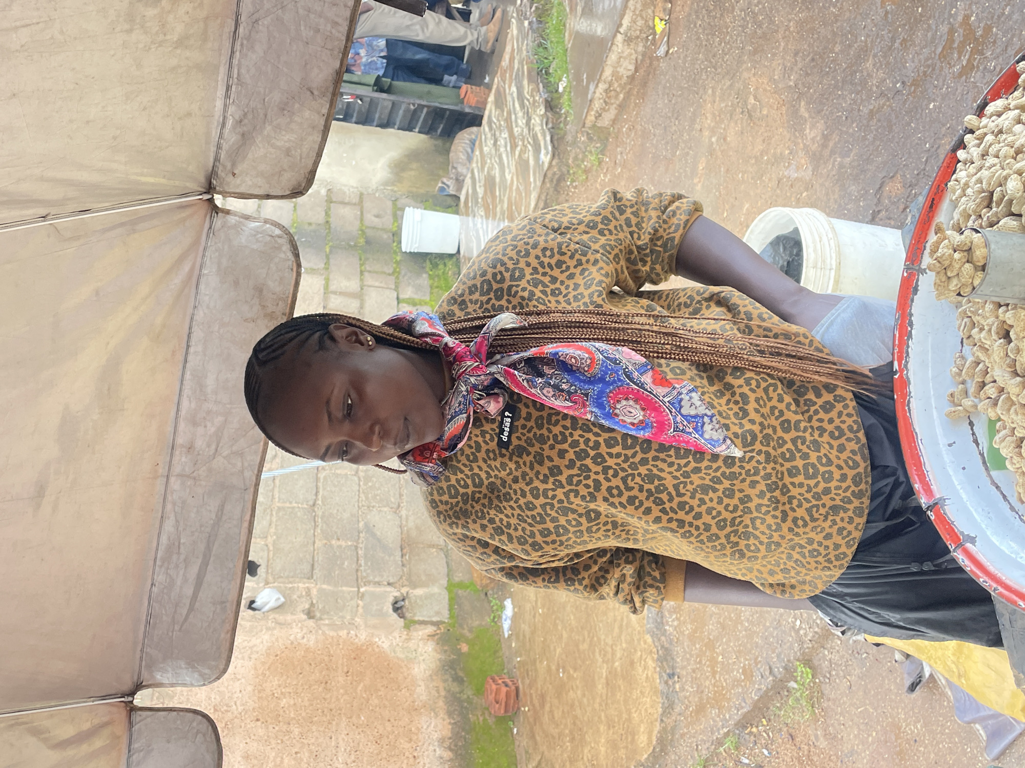 A young Black woman is standing under a beige awning, which is supported by poles. She is wearing a leopard-print sweatshirt, black pants, and a colorful scarf around her neck. Her hair is braided and hangs down her back. In front of her is a large metal bowl filled with peanuts. The background shows a brick wall, a white bucket, and some people walking in the distance. The ground appears to be wet, suggesting it might be raining. The scene appears to be an outdoor market stall in the city of Jos, Nigeria.