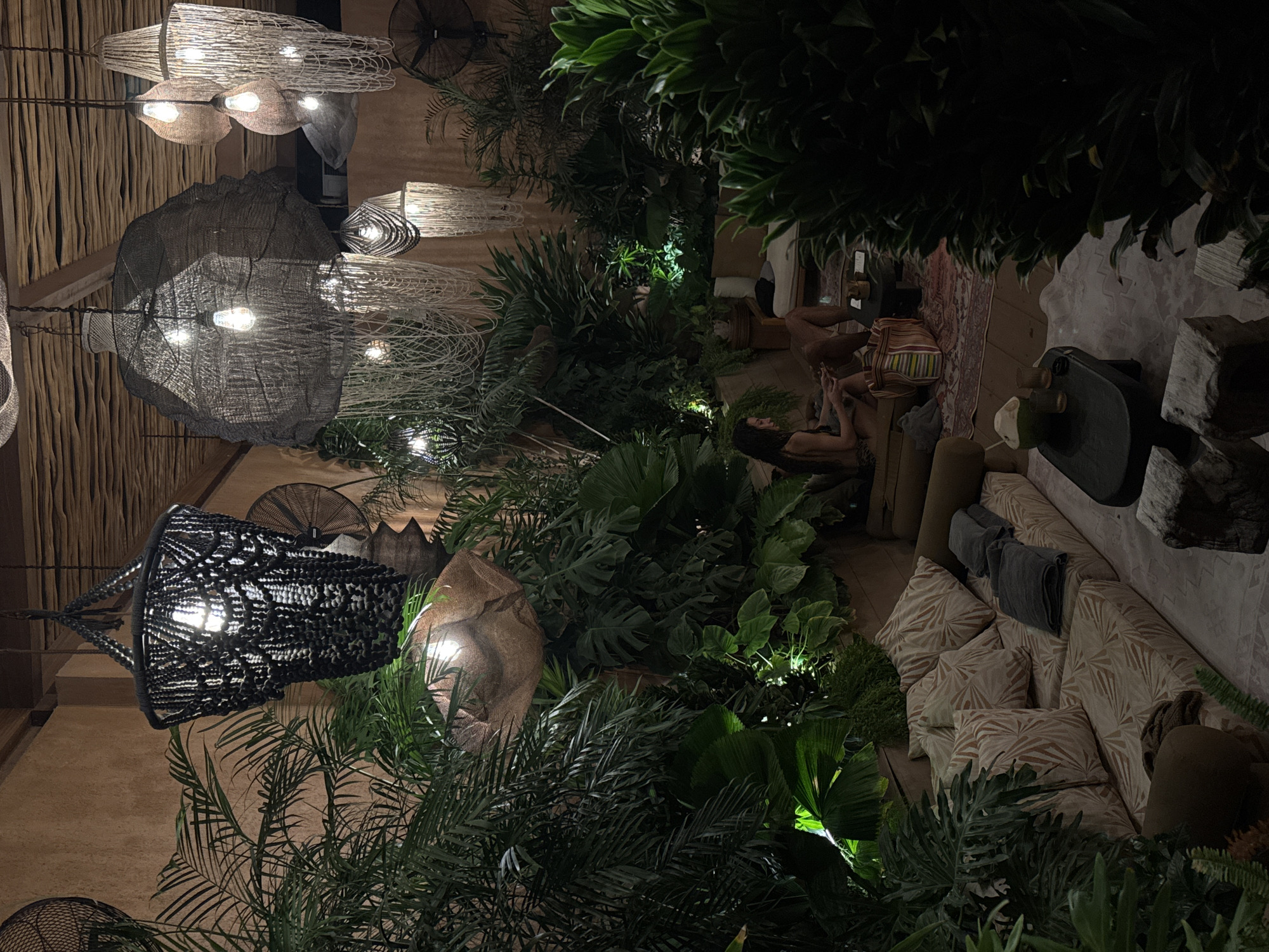 This image captures a dimly lit, tropical-themed lounge area, likely an interior or semi-outdoor space in Kuta Selatan, Indonesia. The environment is characterized by abundant, dense green foliage, including various types of palm leaves and large-leafed plants, creating a jungle-like atmosphere. The walls are textured, light brown or tan, and the ceiling features a dark, slatted wooden structure.
Multiple hanging pendant lights illuminate the scene with warm, ambient light. In the upper left, a large, black, woven or macrame chandelier with exposed bulbs is prominent. To the right, several other large, organically shaped pendant lights made from woven natural fibers (possibly wicker, rattan, or coarse fabric) hang from the ceiling, varying in size and design, some featuring cascading strands. All contain visible Edison-style light bulbs. Several black industrial-style wall fans are visible in the background, mounted on the textured walls.
In the lower-middle portion of the image, two individuals are seated on low cushions in a lounge area. The person on the left is a female with long dark hair, wearing a dark top, seated with bent knees and looking down at an object in her hands. To her right, a second person, appearing male, is partially visible from the lower torso down, wearing dark shorts and seated on the floor.
The seating arrangement consists of a low sofa with light-colored cushions patterned with brown or orange leaf designs. Dark grey folded towels are placed on the sofa. A patterned rug in shades of red and beige with intricate designs covers the floor beneath the seating. A striped fabric tote bag with vertical bands of red, green, blue, and yellow rests on the rug near the seated individuals.
Two small, dark, low-set tables are visible. One, positioned near the seated individuals, holds drinks. Another, in the foreground, displays a green coconut drink with a straw and two glasses. Rough-hewn wooden blocks are present in the immediate foreground, possibly serving as low stools or decorative elements. The overall ambiance is relaxed and tropical.