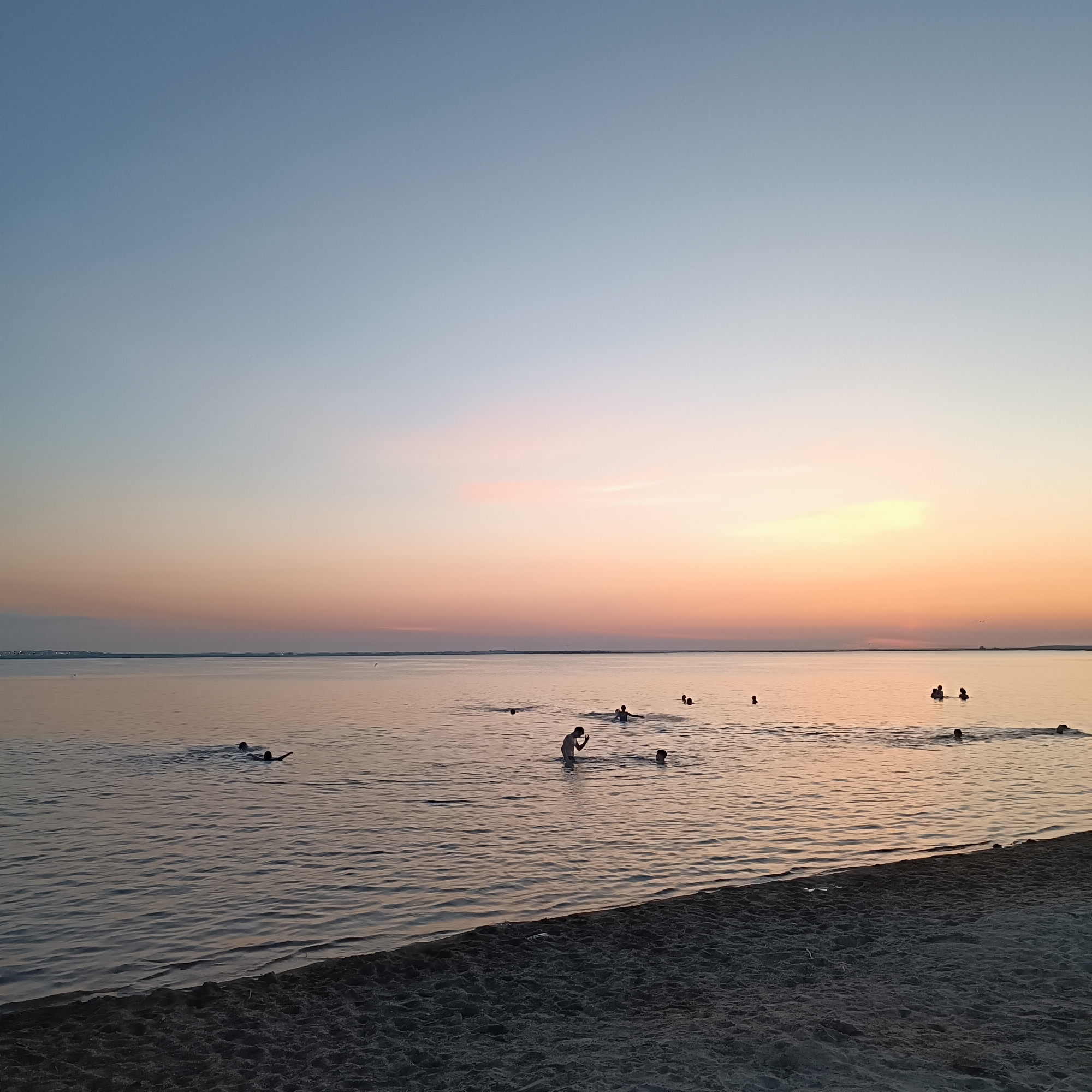 The image captures a serene scene at a beach, likely in Kokshetau, Kazakhstan, during what appears to be twilight. The sky transitions from a pale blue at the top to soft hues of pink and orange closer to the horizon, indicative of sunset or sunrise. A large body of calm water dominates the lower two-thirds of the frame, with the sandy shore occupying the bottom portion. Several individuals, silhouetted against the water, are engaged in swimming or wading. The scene is tranquil, with the gentle ripples on the water reflecting the muted colors of the sky. There are no structures or prominent landmarks visible that would definitively place the location within Kokshetau, but the natural environment suggests a lakeside or coastal setting common in the region.