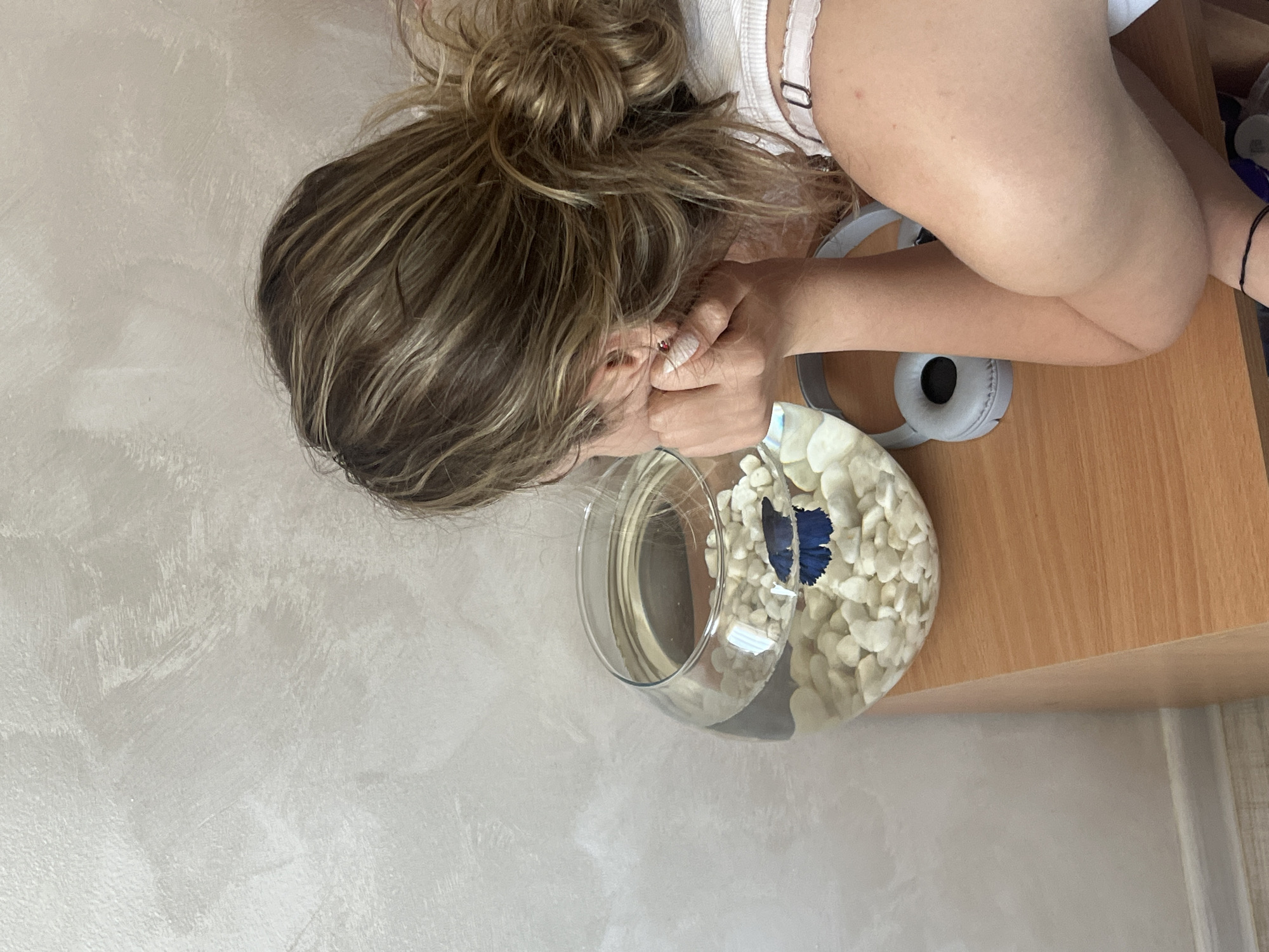 A woman with light brown hair in a messy bun leans over a small fishbowl on a light brown surface. The fishbowl is filled with water and white pebbles, and a blue fish is visible inside. The woman's face is partially obscured by her hands, and she appears to be looking into the fishbowl. Gray headphones are near the fishbowl. The background consists of a light beige wall. The location in the city of Blagoevgrad, Bulgaria cannot be determined from the given information.