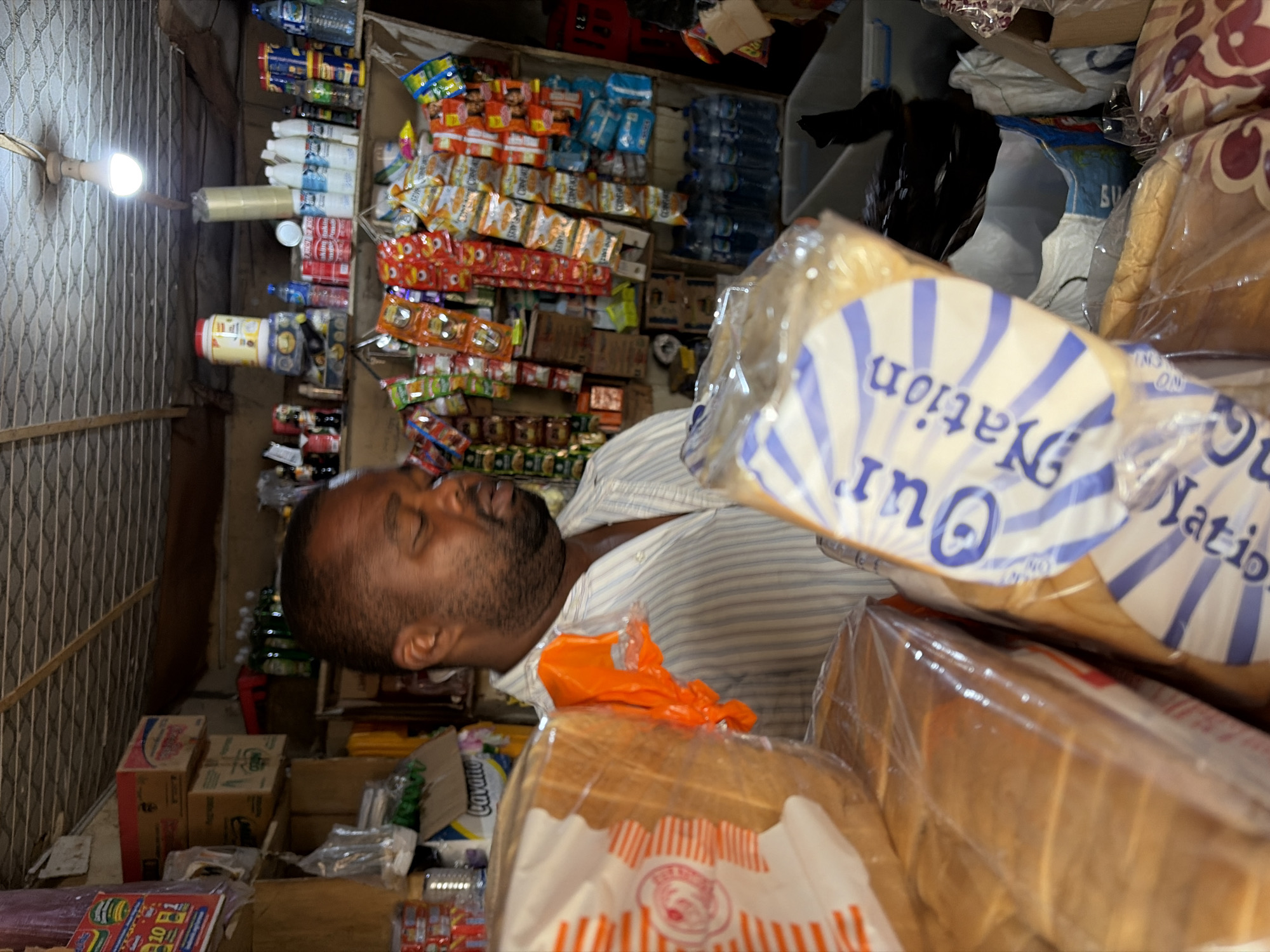 The image captures an intimate scene inside a small, densely packed retail shop, likely a provision store, in Jos, Nigeria. The primary subject is a man, possibly the shopkeeper, who appears to be sleeping or resting with his eyes closed. He has a beard and is wearing a light-colored, striped shirt. He is positioned in the middle-left of the frame, lying on his side or reclining amidst the store's inventory.
The shop is filled with a wide variety of goods, stacked on shelves and in boxes. In the foreground, prominently displayed, are several loaves of packaged bread. The packaging of one loaf clearly shows the brand name "Our Nation" in a blue and white radial design, while another visible loaf has a red and white striped pattern.
Behind the man and the bread, shelves are crammed with numerous small packets of snacks, such as chips or biscuits, and various bottled drinks, including water. Other items like jars, larger containers (one appears to be white and red), and cardboard boxes are also visible, contributing to the cluttered but well-stocked appearance of the store. An orange plastic bag rests on a stack of bread near the man's chest.
A bare light bulb, providing artificial illumination, hangs from a cord on the far left wall, which features a light-colored, textured or tiled pattern. The overall impression is one of a busy, localized business environment where the shopkeeper might snatch moments of rest between customers or during quiet periods. The time of day is not explicitly clear, but the reliance on the single bulb suggests it could be evening, night, or simply a dimly lit interior. The setting strongly indicates a typical small-scale commerce establishment common in Nigerian communities.