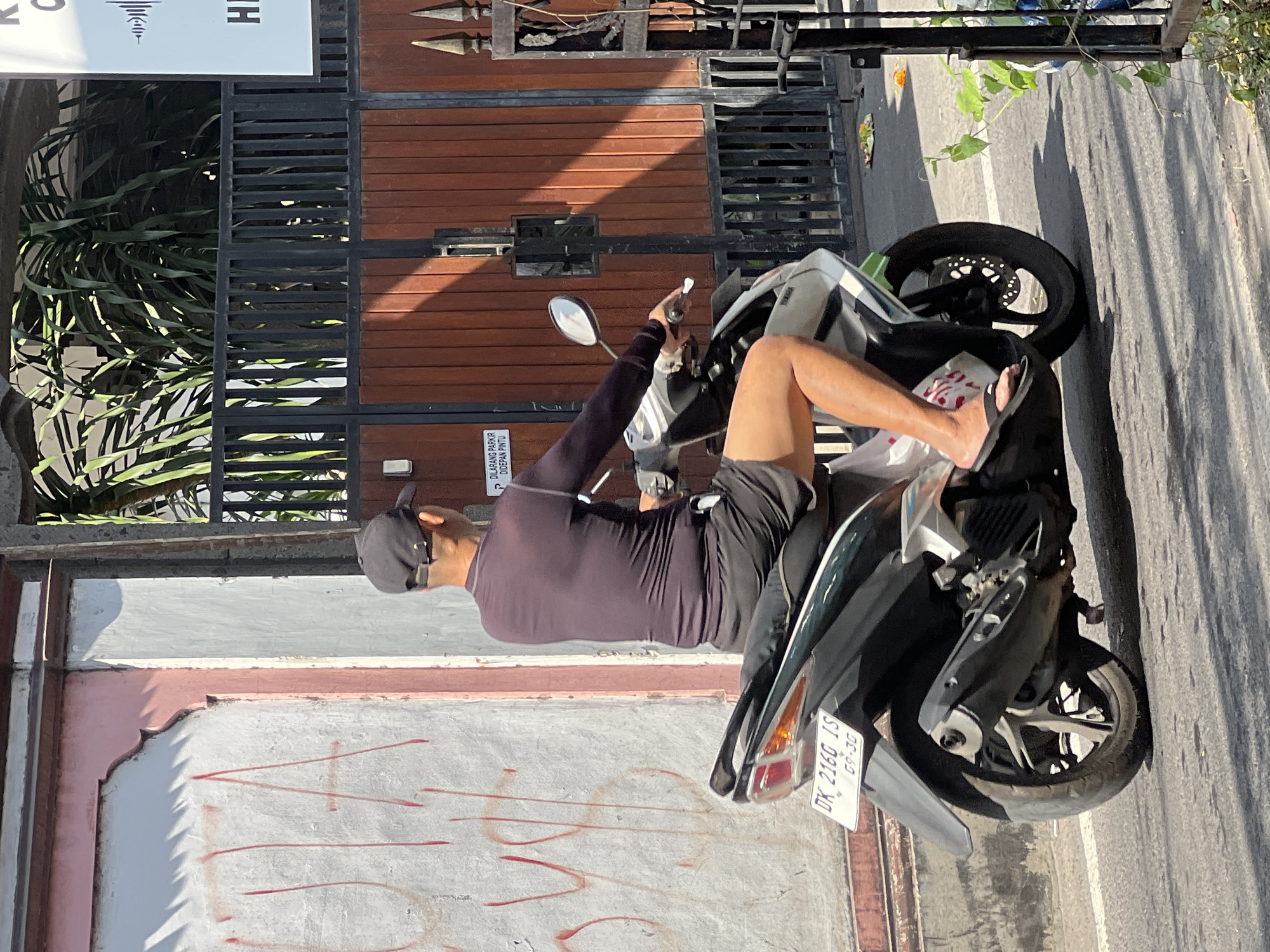 A man wearing a dark long-sleeved shirt, black shorts, a black baseball cap, and black flip-flops is riding a white and black scooter down a paved street in Kuta, Indonesia. The scooter has a white license plate with black lettering that reads "DK 216G IS 09.30". To the left of the street is a weathered, pink and white wall with red graffiti. Behind the man and scooter is a tall wooden gate with black metal bars. The gate is set within a fenced area with foliage visible. Sunlight casts shadows across the scene.