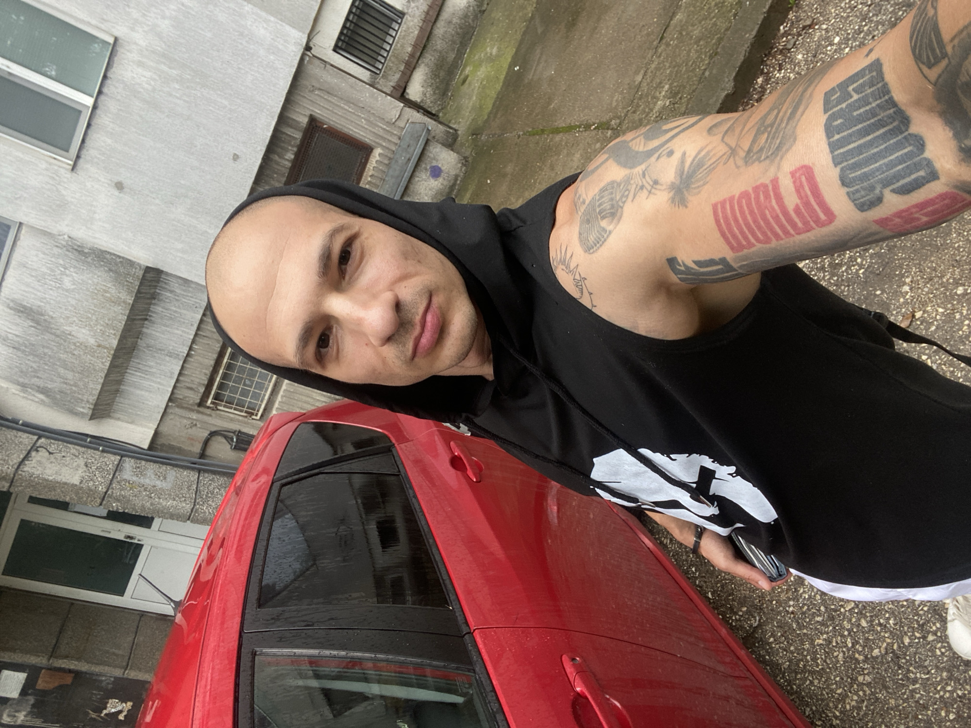 The image captures a selfie of a man outdoors, taken from a slightly high angle looking downwards and to the side. The man has a shaved head and a neutral expression, looking directly at the camera. He is wearing a black sleeveless hoodie with a white graphic print on the left side of his chest. His left arm is heavily tattooed, featuring various designs.
Prominently visible tattoos on his bicep include large red block letters spelling "WORLD," and above it, large black block letters that appear to spell "EMPIRE." Other tattoos on his arm are abstract or stylized, with lines and shapes.
The man is positioned next to a red car, which occupies a significant portion of the lower frame. The car appears damp, suggesting recent rain. In the background, there is a multi-story building constructed of grey concrete or stucco, featuring several windows, some with security bars. The ground is a mix of wet concrete and gravel, with some patches of moss, indicating an outdoor, urban setting in Pleven, Bulgaria. The overall lighting suggests an overcast daytime.