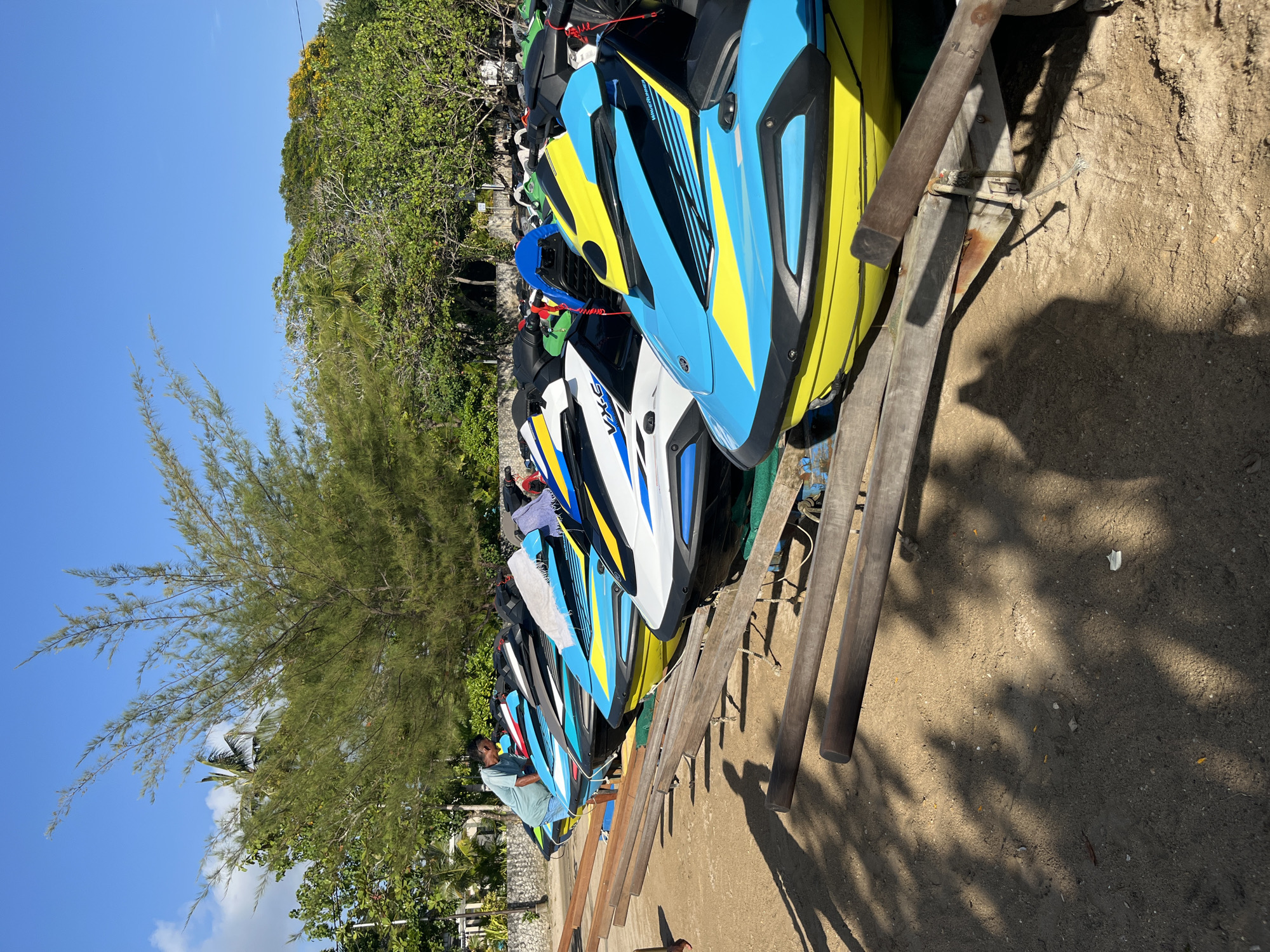 In Kuta Selatan, Indonesia, a line of colorful jet skis are parked on a sandy shore. The jet skis are predominantly yellow, blue, and white with black accents and appear to be new or well-maintained, some with white towels draped over their seats. A man wearing a light blue t-shirt and shorts is standing next to one of the jet skis, appearing to check on it. The scene is set against a backdrop of lush green trees and a clear blue sky, suggesting a tropical or coastal environment. Wooden beams and metal structures are visible, likely used to support and store the jet skis. Shadows of trees and possibly people are cast on the sand, indicating a sunny day.