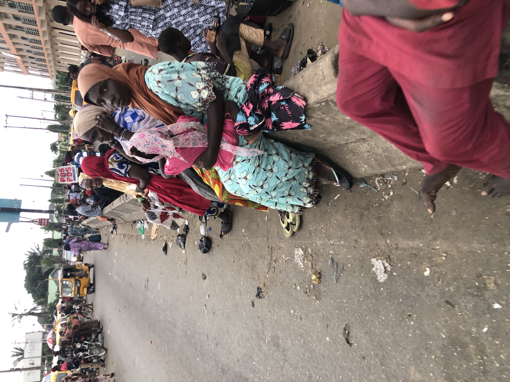 This image depicts a street scene in Ikeja, Nigeria. A group of people, predominantly women, are seated on a raised concrete curb, waiting for transportation. They are dressed in colorful attire, with some wearing headscarves. Various vehicles, including a yellow tricycle (keke napep) and motorcycles, are visible in the background, suggesting a busy thoroughfare. The ground is paved with asphalt and is littered with debris, including plastic bags and discarded items. A multi-story building with arched windows stands to the right of the frame, indicating an urban setting. The sky is overcast, casting a diffused light over the scene.