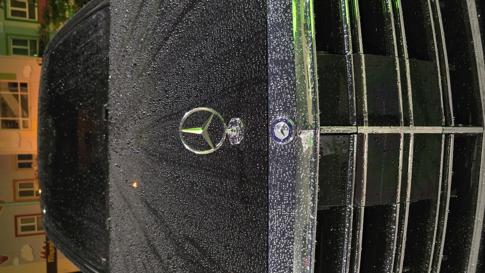 The image displays a close-up, vertically oriented shot of the front section of a dark-colored Mercedes-Benz car, covered extensively with water droplets. The surface appears wet and reflective, suggesting recent rain or a damp environment.
Prominently featured on the car's hood, slightly left of the center, is the iconic silver Mercedes-Benz three-pointed star logo enclosed in a circle. Adjacent to it, to the right, is a smaller, circular metallic badge with a blue center and an indistinct emblem, likely another Mercedes-Benz marque or series badge. The car's grille occupies the right side of the frame, consisting of vertical dark slats with reflective chrome or silver trim, all similarly adorned with water droplets. A striking green light source or reflection is visible on parts of the grille and the wet car surface, adding a distinct hue to the scene.
The background is softly blurred but reveals architectural elements, including colorful buildings with windows, suggesting an urban or residential setting. Faint, warm artificial lights are visible in the background, implying the photo was taken at night or in low-light conditions. There is no clear activity, and the scene is static. No discernible text is visible apart from the Mercedes-Benz logo's design.