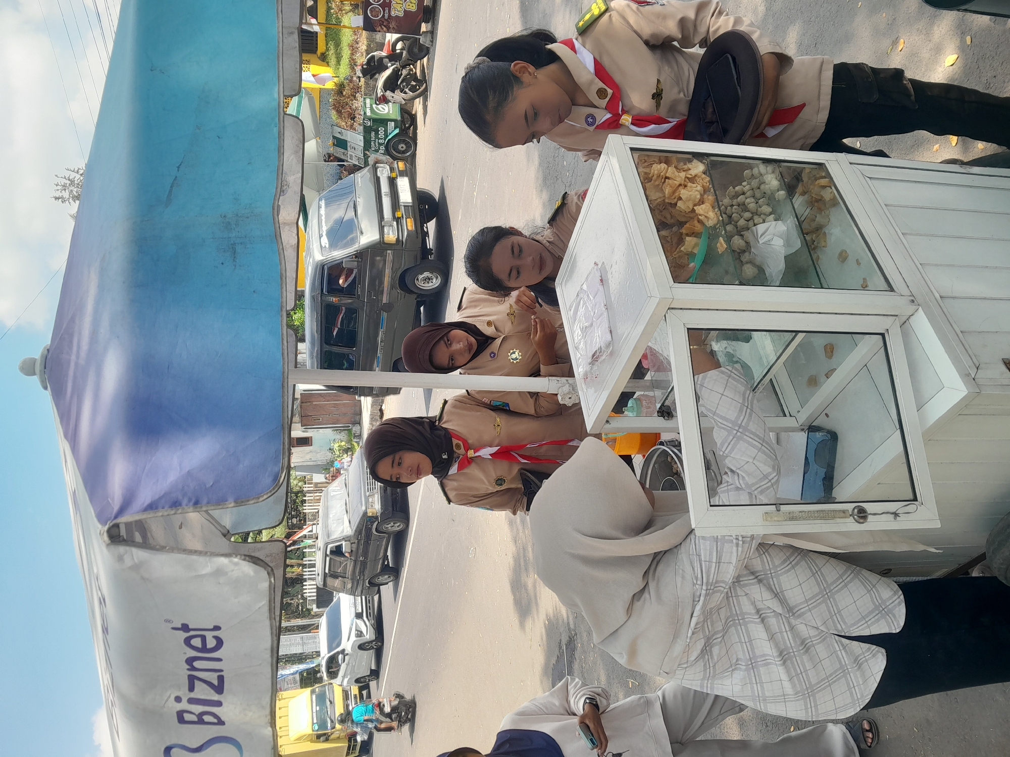 In Genteng, Indonesia, on a bright, sunny day with a clear blue sky, four young women in scout uniforms stand near a mobile food cart. Two of the women are behind the cart, one is leaning over it, and the fourth is to the right, holding a black hat. The food cart is white with glass display cases filled with various food items, including what appear to be snacks and round balls. The women's uniforms are khaki with red and white neckerchiefs and badges. In the background, a street is visible with several vehicles, including a yellow truck, a white car, and a dark-colored van. There are also businesses with signage and a few motorcycles. The scene suggests a street-side vendor operation, likely a business or fundraising activity.