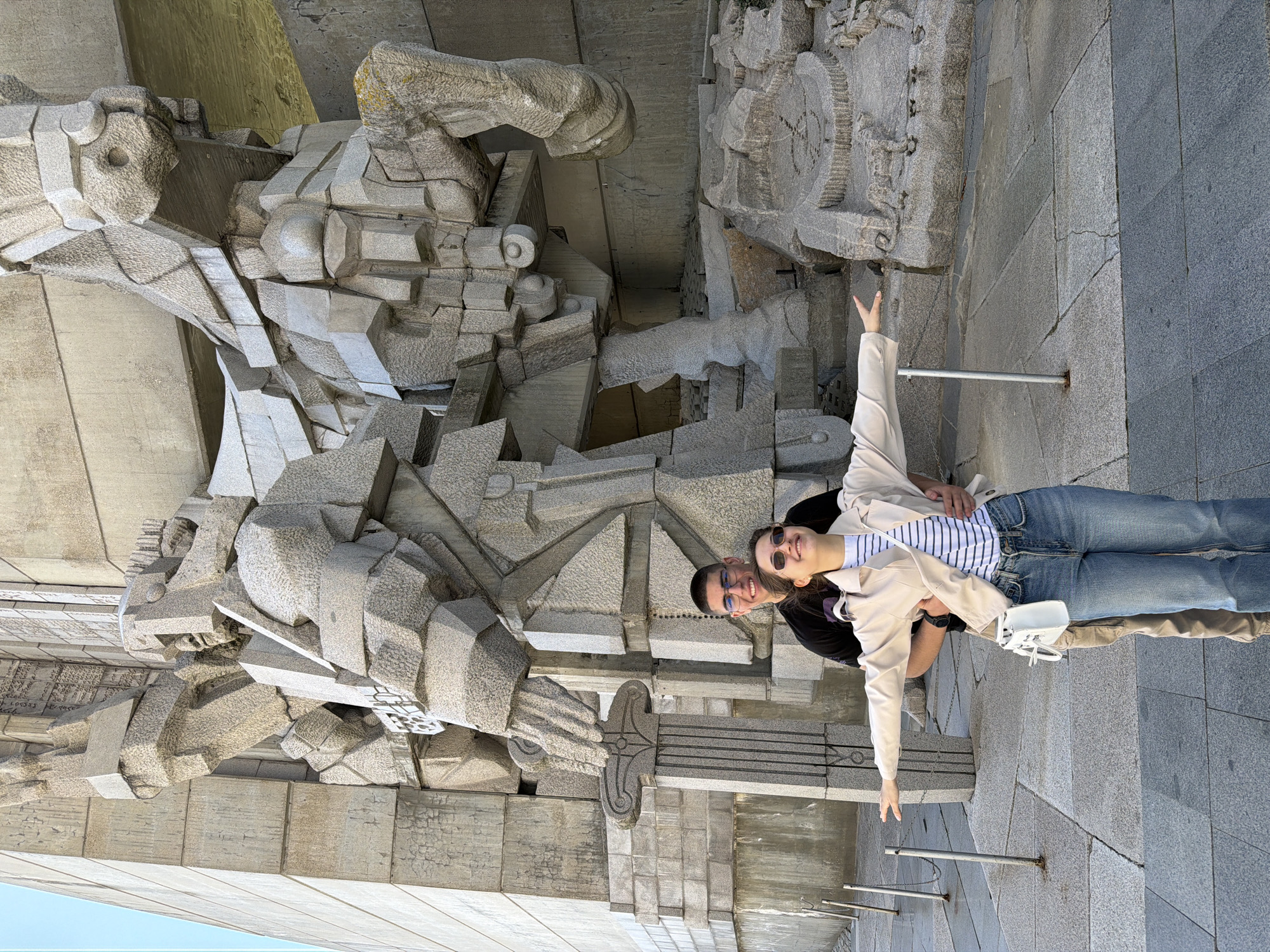 A medium shot shows a couple posing in front of the Monument to the Creators of the Bulgarian State in Shumen, Bulgaria. The large, abstract, concrete sculpture is behind them, occupying most of the left half of the frame. The couple are positioned on stone steps, taking up the right half of the shot. The woman is in the foreground, wearing sunglasses, a striped shirt, blue jeans, and a beige jacket. She has one arm raised. A white purse hangs from her shoulder. The man is behind her, wearing glasses and a black shirt, with one arm around her waist. The steps and a metal railing can be seen in the foreground. The sky is visible in the lower left corner.