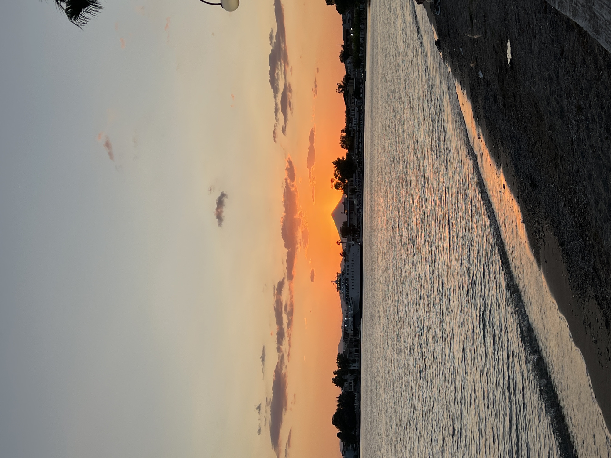 The image captures a serene sunset scene at the port of Eretria, Greece. The sky is painted with warm hues of orange and pink, with scattered clouds adding texture to the twilight canvas. The sun, partially obscured by a distant mountain, casts a golden glow across the water. A large ferry is docked at the harbor, its silhouette visible against the vibrant sky. Along the coastline, buildings and trees form a picturesque backdrop. The foreground features a dark sand beach with gentle waves lapping at the shore, reflecting the colors of the sunset. The scene is peaceful and evokes a sense of tranquility.