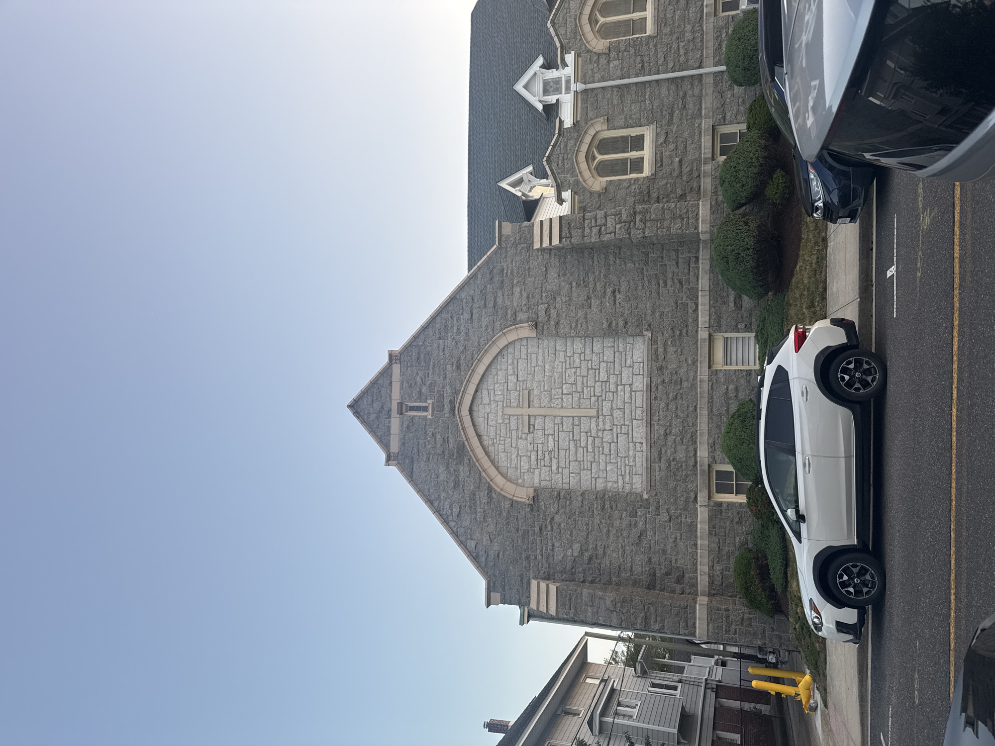 A low-angle, eye-level, full exterior shot captures the facade of a stone church. The church, constructed with rough-hewn, gray granite blocks, features a prominent, arched window with a light-colored stone frame and a subtle cross motif etched into its stonework. Several smaller, arched windows with light-colored stone accents are visible on the lower portion of the building. The roof is dark gray asphalt shingles. Dormer windows with white trim project from the roof. Adjacent to the church is a two-story residential building with gray siding and white trim. In the foreground, a white SUV is parked at the curb. Further down the street, a dark-colored SUV is also parked. A yellow traffic barrier stands on the sidewalk near the church. The sky is a clear, pale blue. The scene is a street in Ocean City, United States of America.