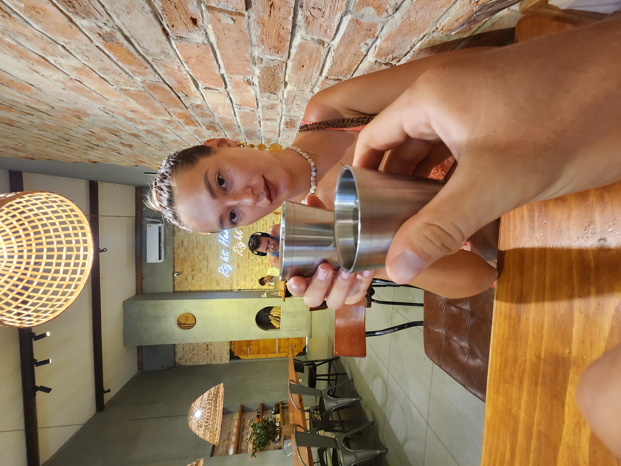 A close-up, eye-level shot captures a couple toasting with small metal cups in what appears to be a cafe or restaurant in Da Nang, Vietnam. The woman, in the foreground, is holding one cup and looking at the camera with a slight smile. She has her hair tied up in a bun and is wearing earrings and a necklace. The man's hand, in the foreground, is holding the other cup, obscuring his face. In the background, another person can be seen wearing headphones and working on a laptop. The cafe has a rustic ambiance with exposed brick walls, wooden furniture, and woven light fixtures. The overall scene suggests a casual and intimate moment shared between the couple.