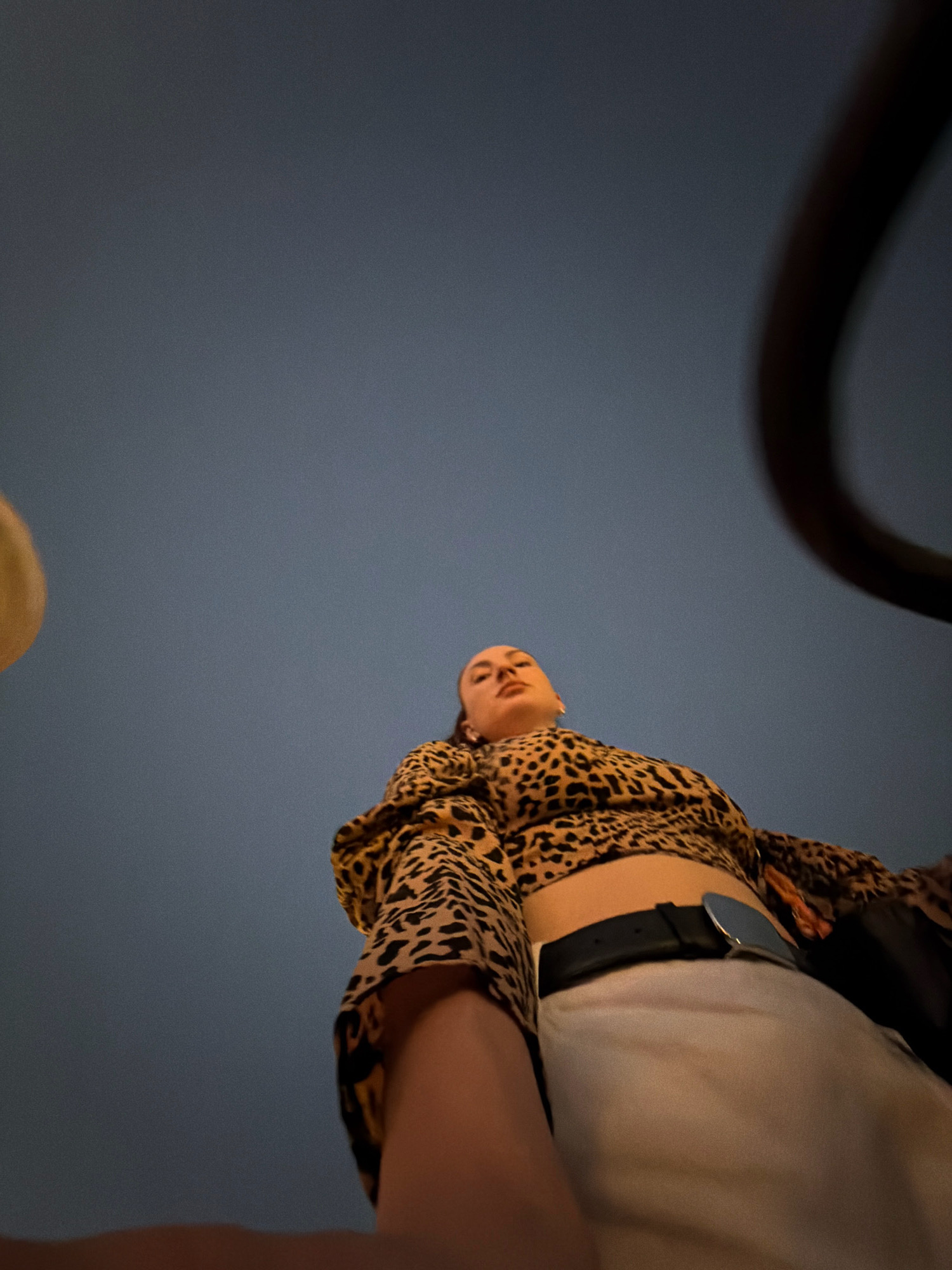 A low-angle, full-body shot captures a woman standing outdoors at dusk in Sharm El Sheikh, Egypt. She is wearing a leopard-print top, a white skirt, and a black belt. Her head is tilted back and she is looking upwards. The sky is a deep blue, transitioning to a darker shade at the top of the frame. A dark, curved object, possibly part of a structure or vehicle, is visible in the upper right corner. A blurry, yellowish circular object is present in the upper left corner. The overall lighting suggests an outdoor setting at twilight.