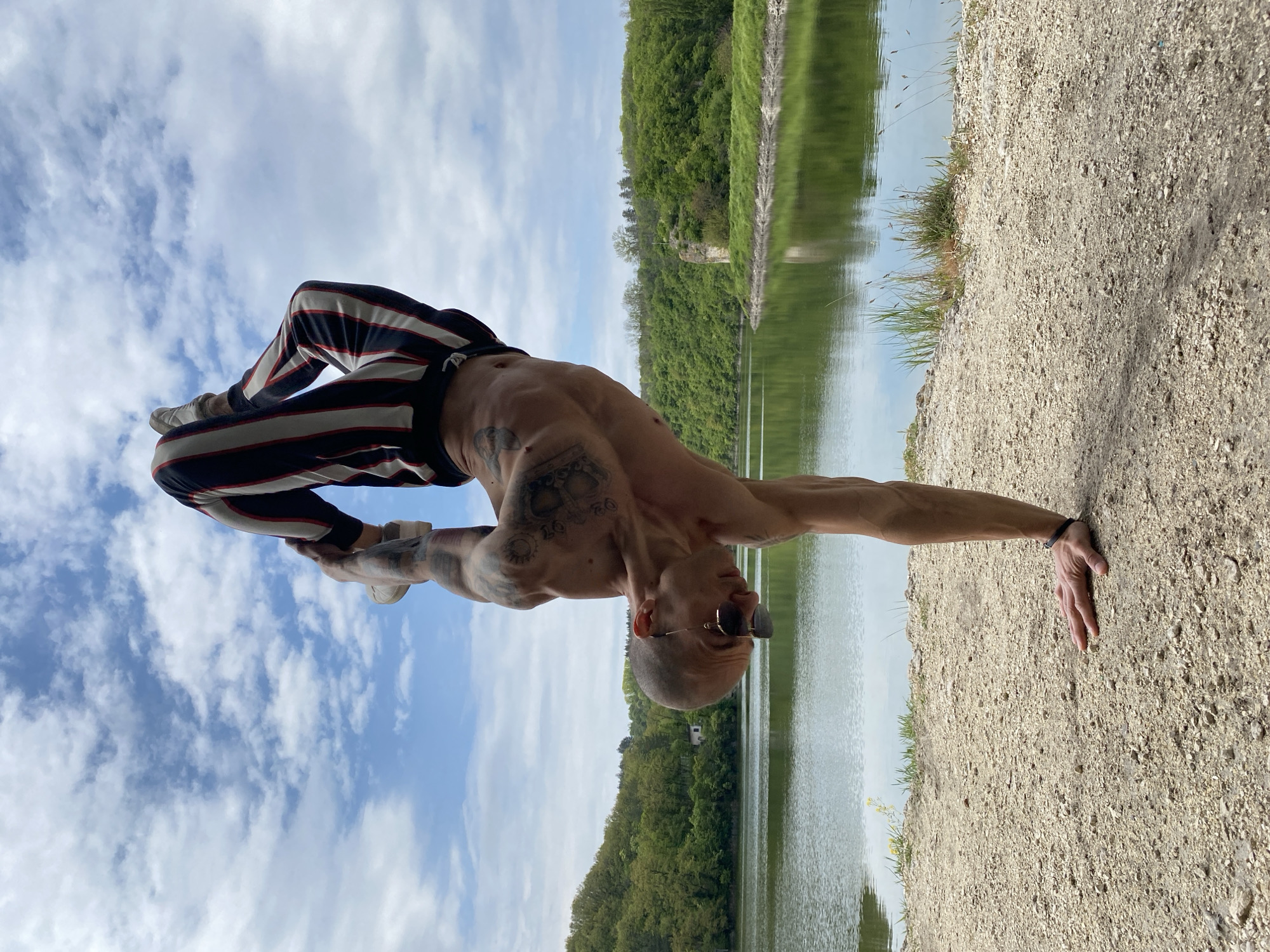 A man is captured in a dynamic yoga pose outdoors near Kaylaka Park, Pleven, Bulgaria. He stands on one hand on a gravel path beside a lake. His body is curved, legs are bent, and he is wearing sneakers and vertically striped pants. He is bare-chested, and tattoos are visible on his upper torso and arms. The background includes the lake, trees, and a partially cloudy sky.