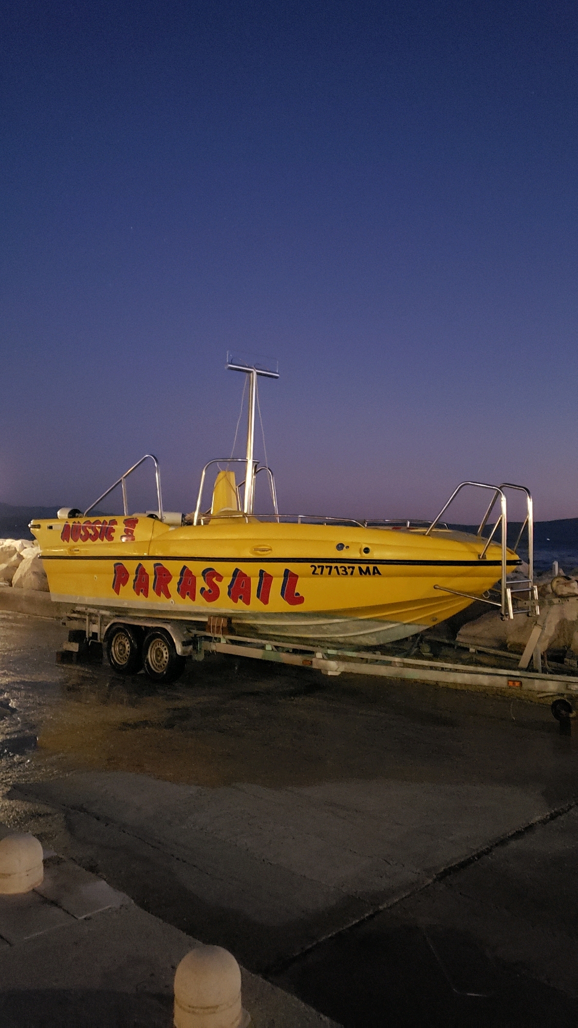 In Općina Tučepi, Croatia, a yellow boat named "AUSSIE II PARASAIL" with registration number "277137 MA" is parked on a trailer at what appears to be a waterfront. The boat is positioned on a paved surface with some scattered rocks and white bollards visible in the foreground. The sky is a deep blue, suggesting it is dusk or dawn, and the sea is visible in the background. The scene is illuminated by artificial light, casting a glow on the boat and surrounding area. There are no people visible in the image.