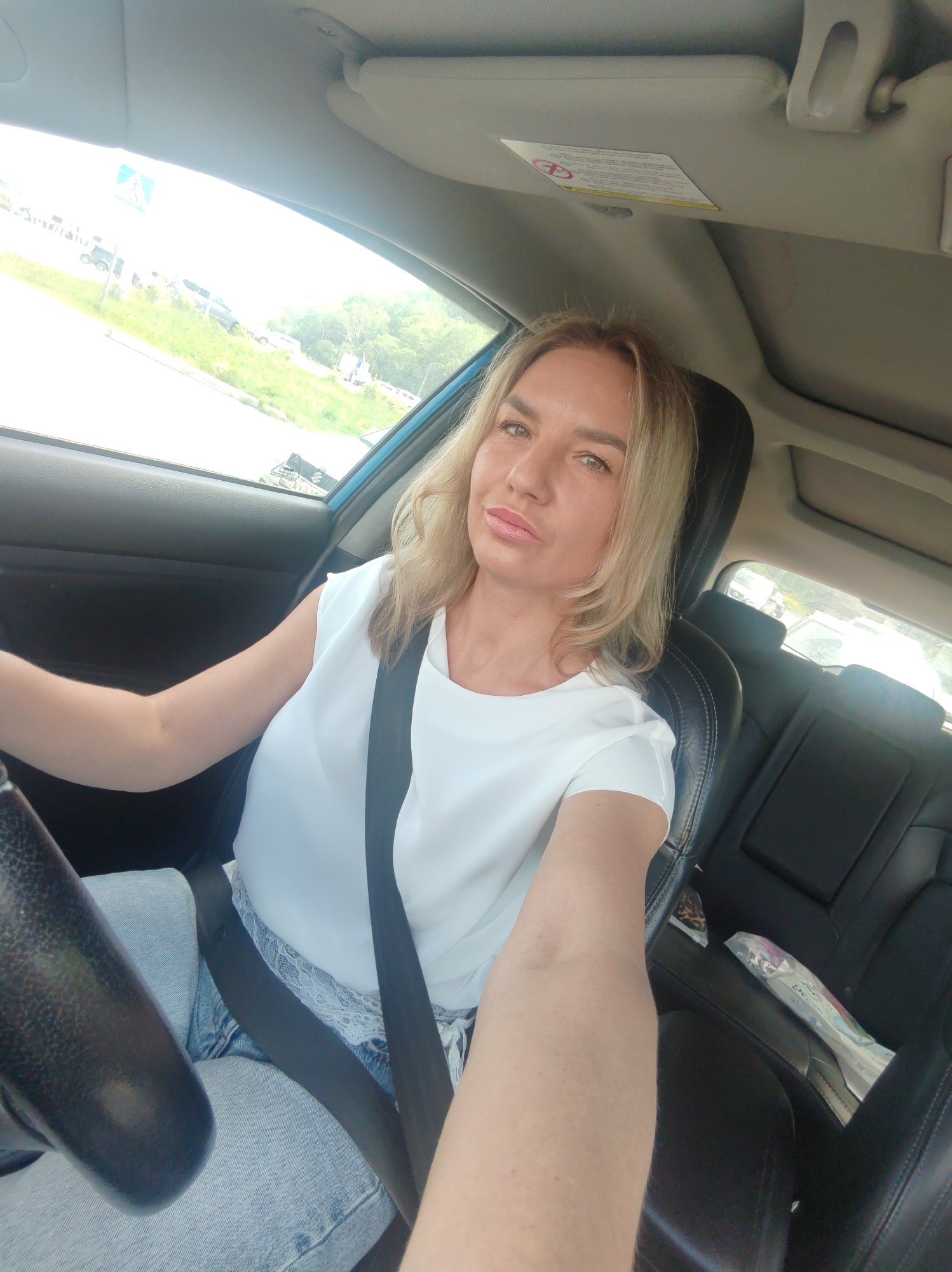 A medium shot captures a blonde woman with her hair down, wearing a white short-sleeved shirt and blue jeans, sitting in the driver's seat of a car. She is looking towards the camera with a neutral expression. Her right arm is on the steering wheel, and her left arm is bent, resting on her lap. A black seatbelt is visible across her chest. The interior of the car, including the dashboard, steering wheel, and backseats, is visible. Through the car window, a blurred view of an outdoor environment, possibly a street with trees and other vehicles, can be seen. The overall lighting suggests it is daytime.