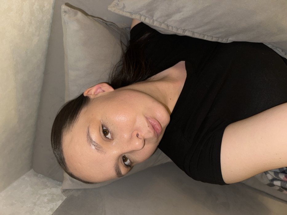 An adult female is depicted in a close-up, semi-reclined position, presumably indoors in the city of Chaikovsky, Russia. She has dark brown hair pulled back, brown eyes, and a light to medium complexion with a natural sheen visible on her forehead and cheeks. Her lips are slightly parted in a subtle pout. She is wearing a black short-sleeved t-shirt. Her head rests on a light grey/beige pillow, and a similarly colored blanket or duvet partially covers her left side. A patterned plaid blanket, featuring light and dark tones, is visible on the lower left of the frame. The immediate background consists of a light grey/off-white textured wall or wallpaper.