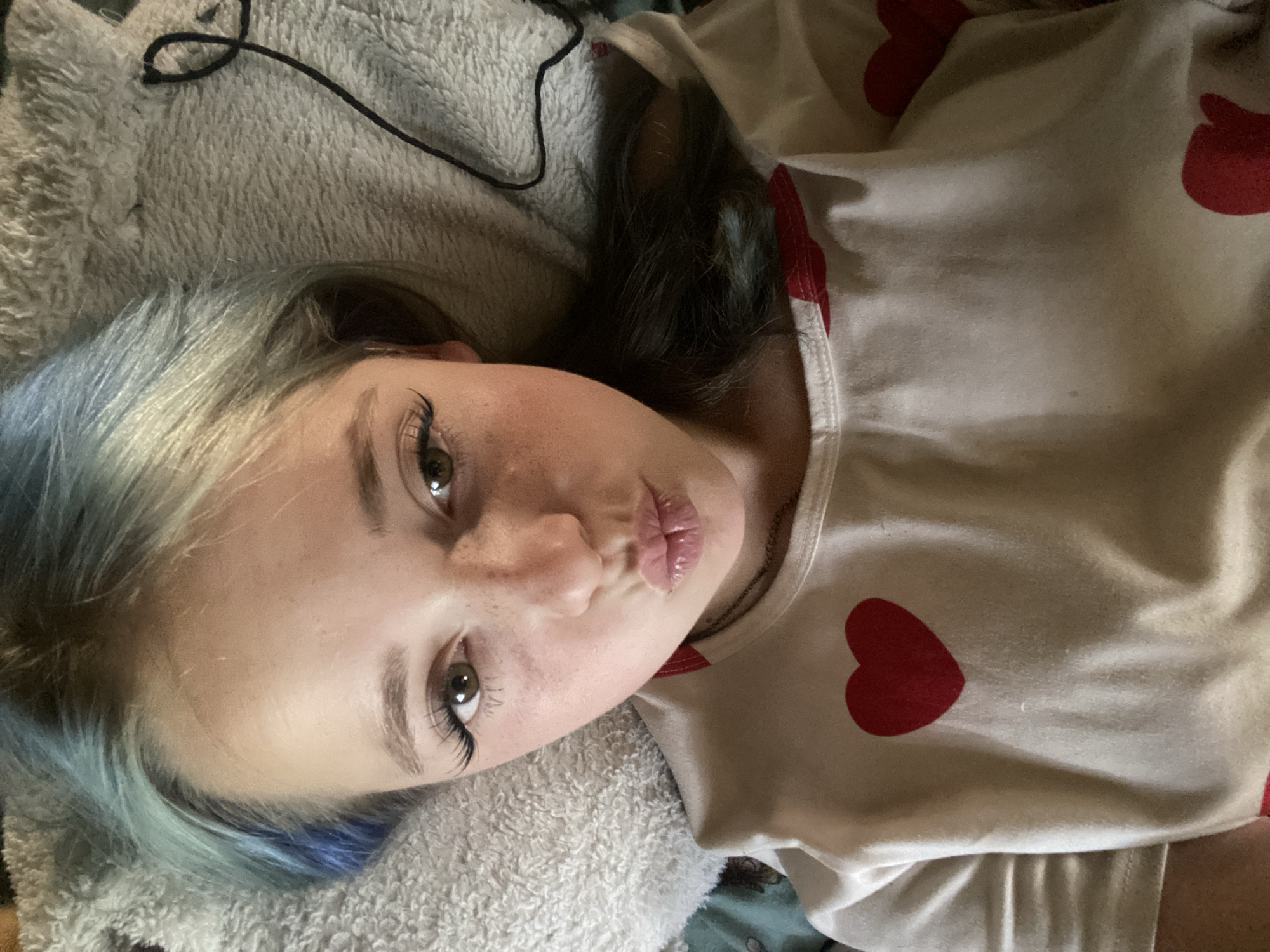 The media file is a close-up, overhead shot of a young person lying on a soft, textured surface. The person has light-colored skin with freckles across their nose and cheeks. Their eyes are open and looking towards the camera, with long, dark eyelashes. Their lips are pursed, as if blowing a kiss. The person has dyed hair with streaks of blue and dark brown. They are wearing a light-colored shirt with a pattern of red hearts. The background consists of a plush, light-colored blanket and a dark cord. The lighting appears to be indoor, with no clear indication of the time of day or weather. The overall emotion conveyed is playful or affectionate. There is no visible text in the media file.
