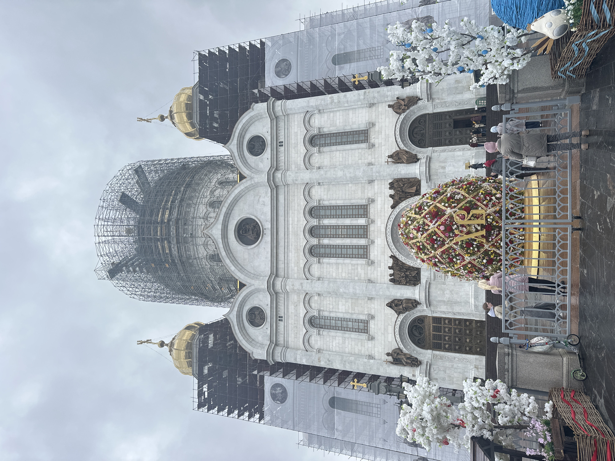 A large, ornate cathedral, identified as the Cathedral of Christ the Savior in Moscow, Russia, stands under an overcast sky. The imposing structure features a bright white stone facade adorned with multiple arched windows, intricate carvings, and numerous bronze sculptures depicting religious figures. Several golden domes crown the building, though a significant portion of the cathedral, most notably the large central dome and at least one smaller adjacent dome, is extensively covered in scaffolding, indicating ongoing renovation or maintenance work. The scaffolding structure is complex and intricate, enveloping the domes.
In the foreground, festive Easter decorations are prominently displayed. A colossal, colorful egg-shaped floral arrangement, richly decorated with various flowers and foliage in shades of red, yellow, and green, sits near the cathedral entrance. The letters "XB," a traditional Russian Orthodox Easter greeting meaning "Christ is Risen," are clearly visible on the side of this floral egg. Nearby, artificial trees with white blossoms and delicate blue egg ornaments contribute to the festive atmosphere. Another wicker-basket-like decoration is also visible.
Several people are gathered near the main entrance of the cathedral, some appearing to queue or observe the decorations and the building. The ground appears damp, suggesting recent rain or simply reflecting the overcast weather conditions. The scene captures a moment of religious observance or sightseeing at this iconic landmark, blending ongoing structural work with a period of celebration, likely Easter. The time of day is daytime, under a uniformly gray, cloudy sky.