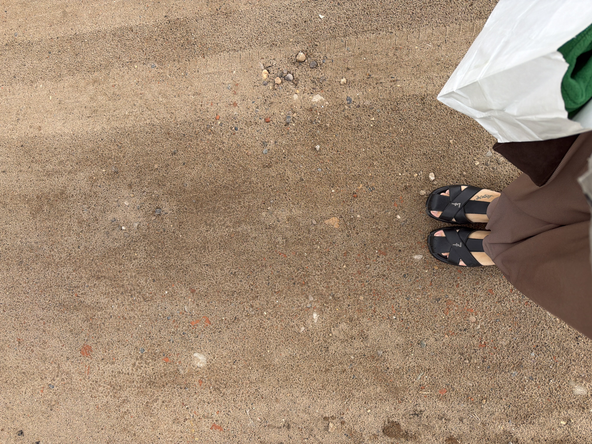 The image shows a top-down view of a person's feet wearing black sandals and brown pants, standing on a sandy or gravelly surface. A white shopping bag is partially visible on the right side of the frame. The ground is textured with small stones and appears to be a pathway or outdoor area. The scene is devoid of any discernible landmarks or specific architectural features that would pinpoint a location within the city of Chaikovsky, Russia.