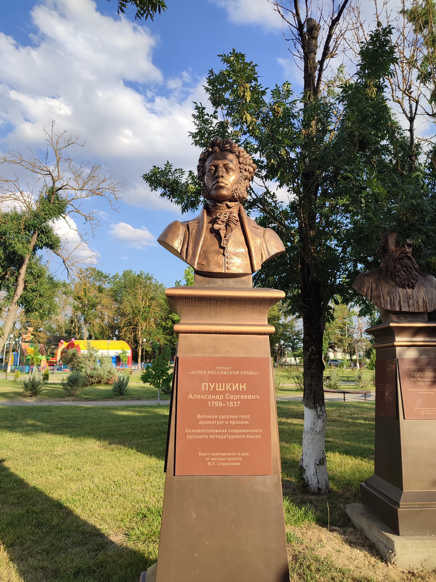 The image shows two bronze busts on pedestals in an outdoor park. The bust in the foreground is of Alexander Pushkin, a renowned Russian poet, playwright, and novelist. The pedestal has an inscription that reads, "PROJEKT <<ALLEYA ROSSIYSKOY SLAVY>> PUSHKIN Aleksandr Sergeevich 1799-1837 Velikiy russkiy poet, dramaturg i prozaik Osnovopolozhnik sovremennogo russkogo literaturnogo yazyka Byost prenesyon v dar ot avtora proekta M. L. Serdyukova." This translates to: "PROJECT <<ALLEYS OF RUSSIAN GLORY>> PUSHKIN Alexander Sergeevich 1799-1837 Great Russian poet, playwright and prose writer Founder of the modern Russian literary language The bust was presented as a gift from the project author M. L. Serdyukov."
To the right of Pushkin's bust, partially visible, is another bust, likely of Leo Tolstoy, given the inscription visible on its pedestal: "PROJEKT <<ALLEYA ROSSIYSKOY SLAVY>> TOLSTOY Lev Nikolaevich 1828-1910 Velikiy russkiy pisatel i myslitel." This translates to: "PROJECT <<ALLEYS OF RUSSIAN GLORY>> TOLSTOY Leo Nikolaevich 1828-1910 Great Russian writer and thinker."
The park setting includes green grass, trees, and a bright blue sky with scattered white clouds. In the background, there are colorful inflatable structures, possibly a playground, and other park amenities like benches and lampposts. The overall scene suggests a public park in the city of Abinsk, Russia, dedicated to honoring prominent figures of Russian culture. There are no people clearly visible in the image.