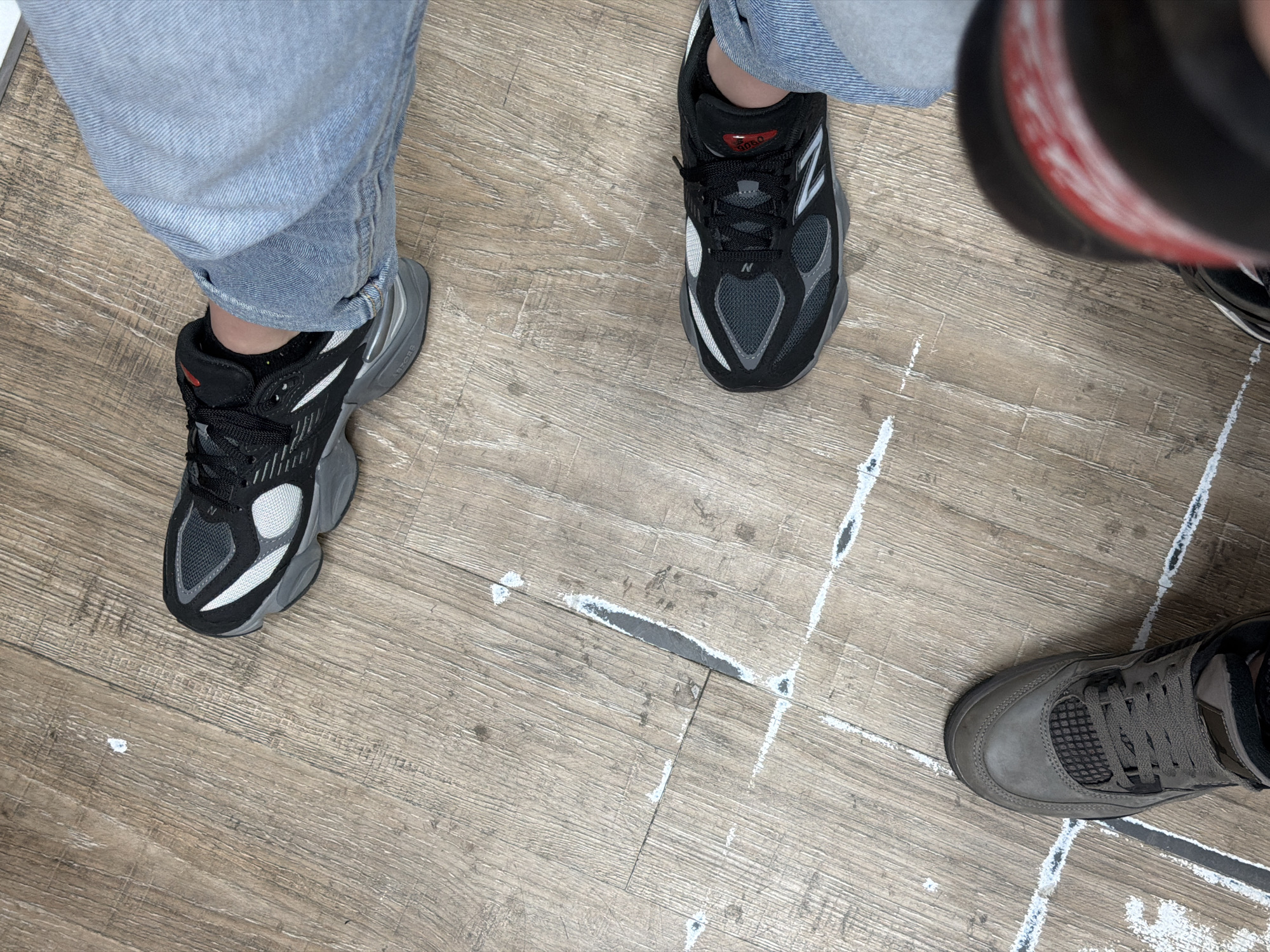 The image shows three pairs of shoes on a light-colored wood floor. Two pairs are black and gray New Balance sneakers, and the third is a gray-brown shoe with laces. The legs of people wearing the shoes are visible, with one pair of legs clad in light-wash denim jeans. White paint or chalk lines are present on the floor. No information suggests a location in Plovdiv, Bulgaria.