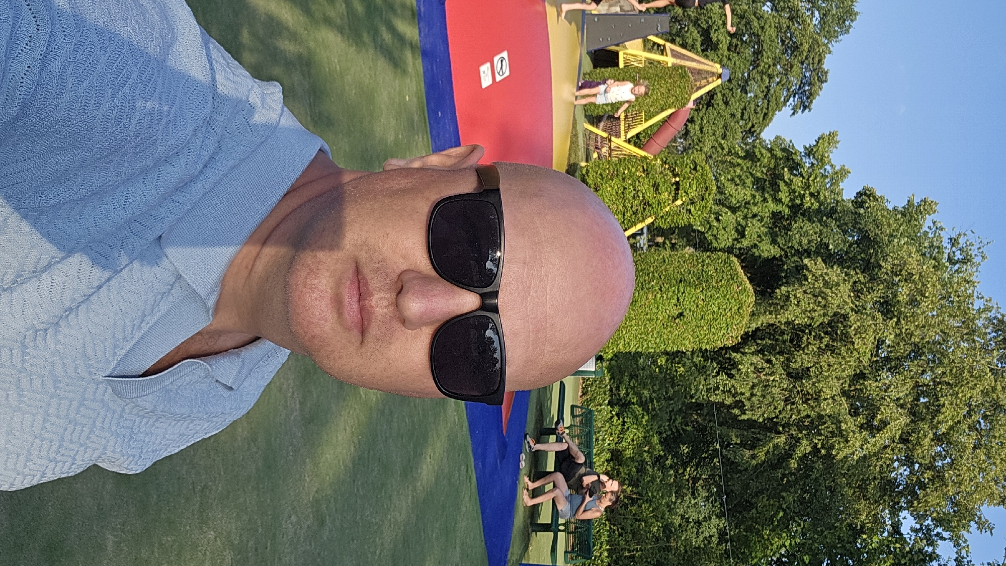 A bald, fair-skinned man wearing sunglasses and a light blue collared shirt is taking a selfie outdoors. In the background, a playground structure with yellow and red components is visible, with a young girl standing near it. Further back, two people are sitting on a green bench. The scene appears to be a park or recreational area in the city of Groenlo, Netherlands, given the presence of a playground and lush greenery. The lighting suggests it is daytime, likely late afternoon or early evening due to the long shadows.