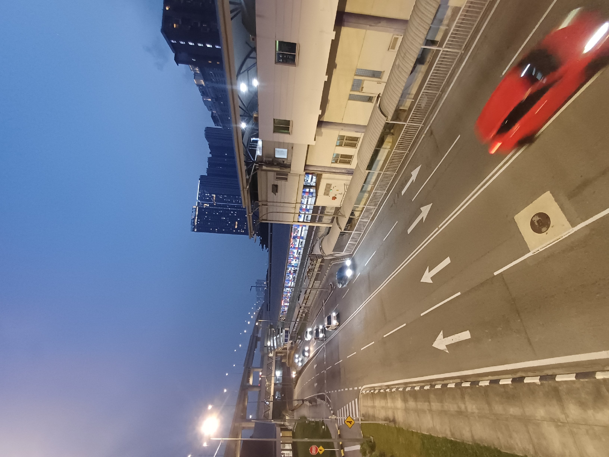 The image captures an elevated view of a road and an adjacent building at dusk in Kuala Lumpur, Malaysia. The sky is a deep blue, suggesting the transition from day to night. Multiple lanes of traffic are visible on the road, with several cars driving in both directions. One car, a red sedan, is in motion, creating a blur due to the long exposure. The building beside the road appears to be a transit station or a commercial establishment, with a covered walkway along its length. Streetlights illuminate the scene, and in the background, tall residential or office buildings are visible, some with their windows lit. A multi-level overpass or flyover is also present, indicating a busy urban environment. The overall atmosphere is one of a modern city in the evening, with ongoing activity on the roads and in the surrounding infrastructure.