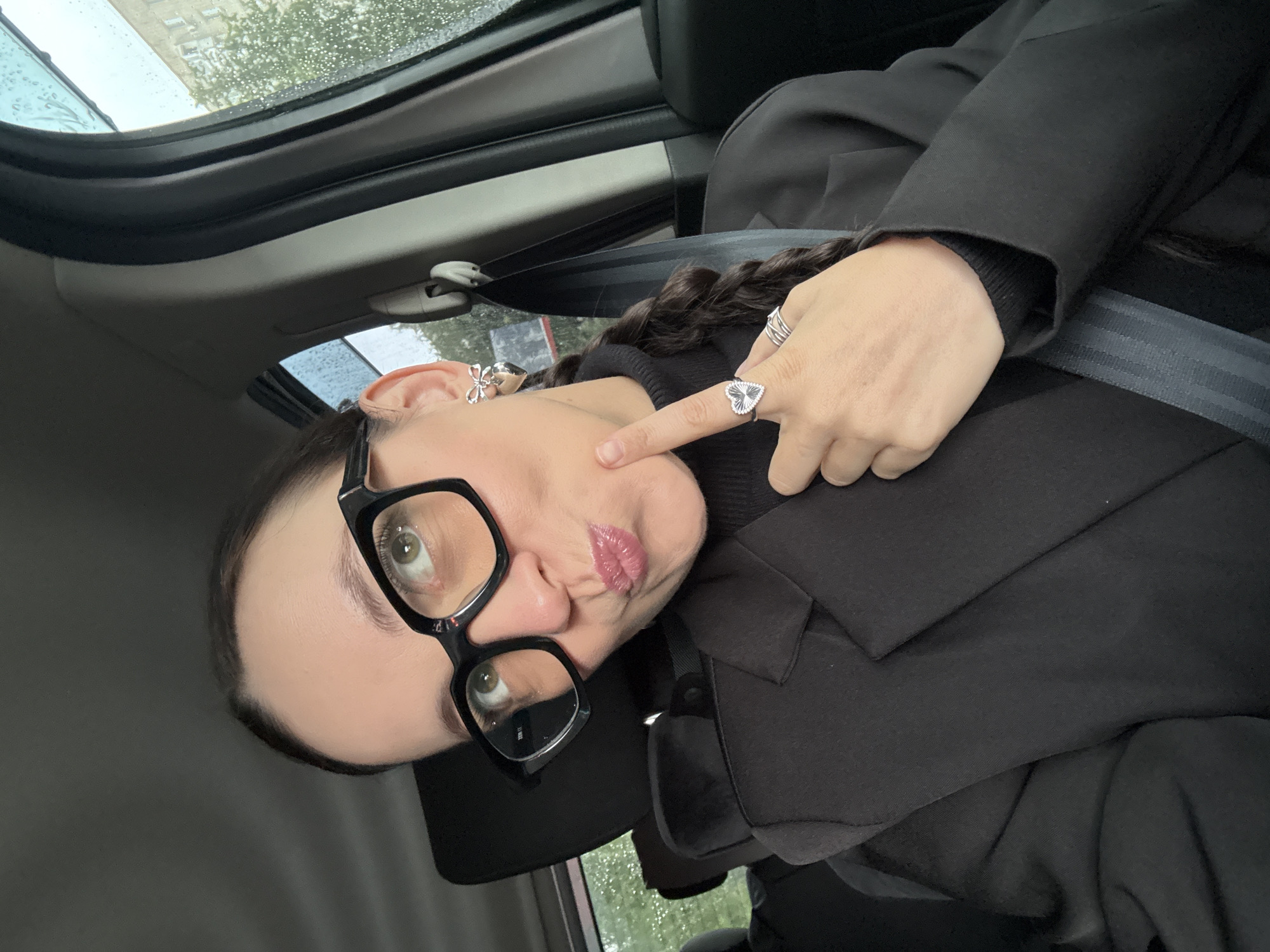 A woman with dark hair and light green eyes is in the passenger seat of a car, looking up and to her left. She is wearing black-rimmed glasses, a black turtleneck, a black blazer, and has a silver heart-shaped ring on her right index finger. Her left ear has a silver bow-shaped earring, and her right ear has a silver heart-shaped earring. Her lips are pursed. Rain is visible on the car window to the right, and a building and trees can be seen through the window. The image was taken in Chaikovsky, Russia.