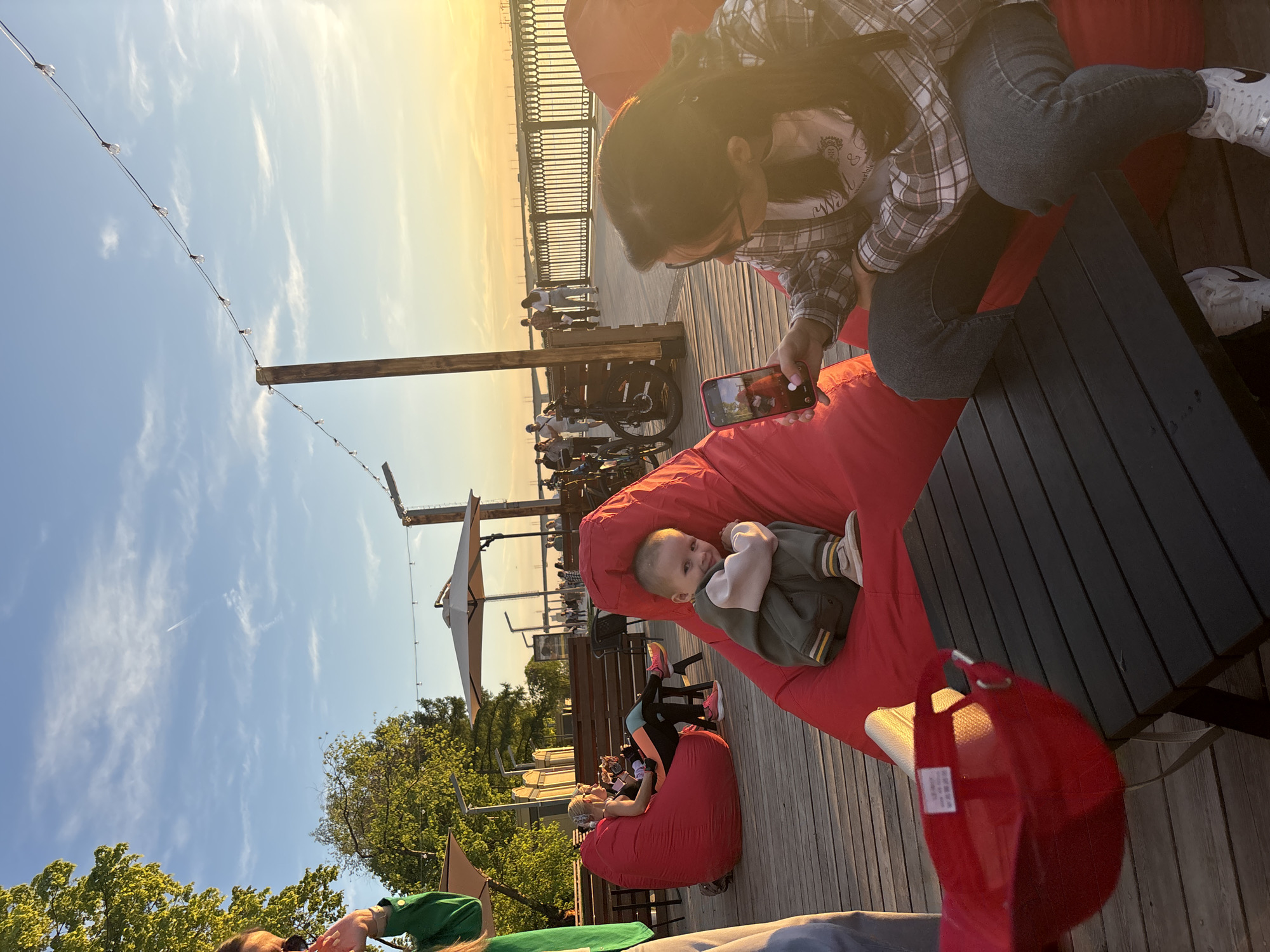 The image depicts an outdoor scene likely taken in the city of Perm, Russia. The setting appears to be a leisure area with wooden decking, possibly a rooftop or elevated platform. People are seated on red beanbag chairs. A woman in a plaid shirt is seen looking at her phone, while a baby sits nearby in another beanbag. In the background, a railing overlooks what seems to be a cityscape during sunset. The sky shows gradient colors of blue to orange, suggesting either sunrise or sunset. String lights are strung overhead.