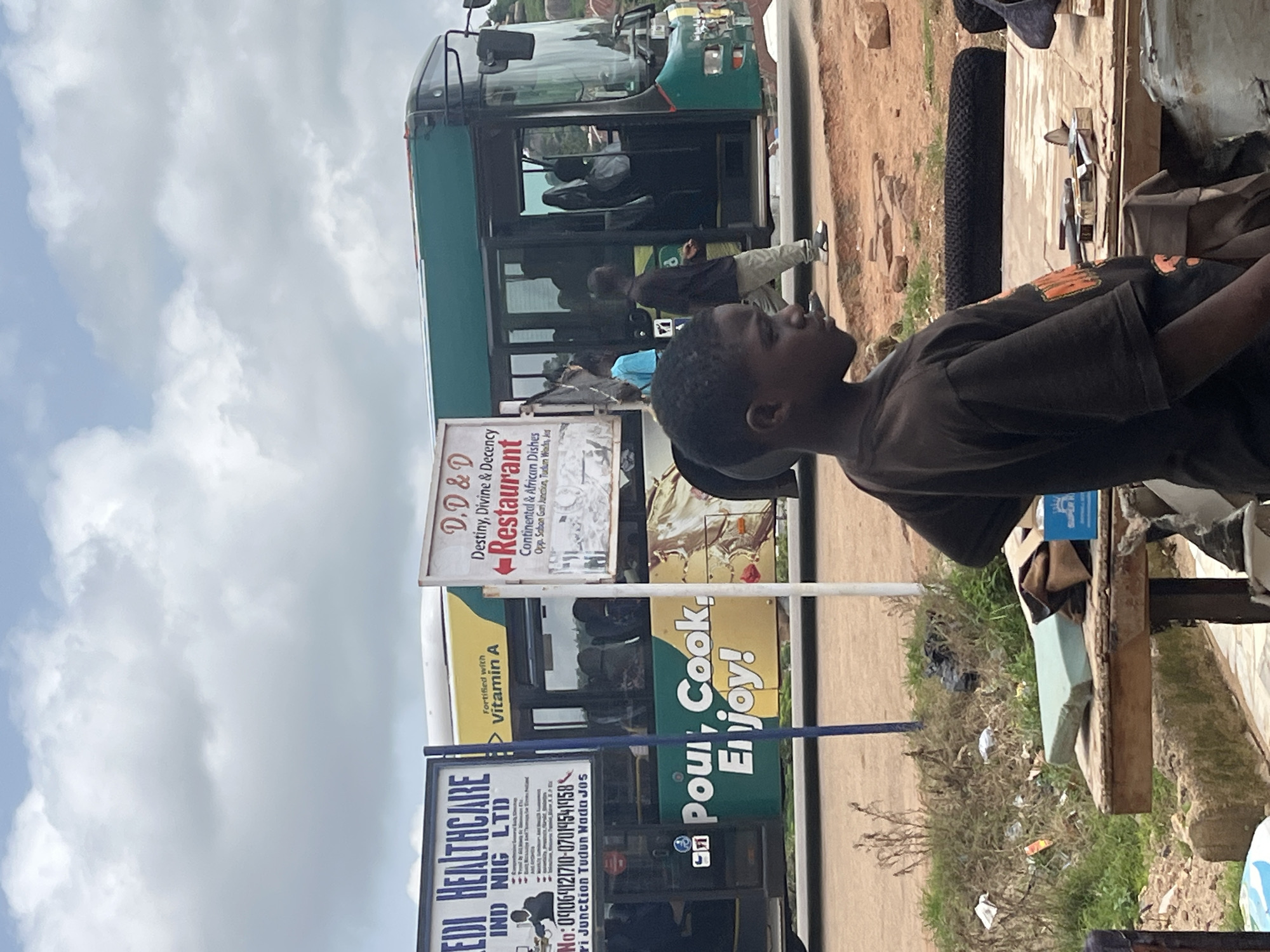 An outdoor medium shot captures a street scene in Jos, Nigeria, featuring a large bus and several individuals. In the foreground, a young boy wearing a dark brown t-shirt is positioned on the right, looking to the left. He is standing behind a wooden table with various items on it, including tools such as a hammer and what appears to be a knife or sharp object, and some fabric. The table is cluttered with debris, and a blue box with white text is visible.
Behind the boy and the table is a green and yellow bus, likely a public transport vehicle. A sign on the side of the bus reads "Pour, Cook, Enjoy!". To the left of the bus, a blue and white sign displays "EDDI HEALTHCARE IND. NIG. LTD." and contact information. Another sign, located above the bus, has the text "D.D & D Restaurant" with directional arrows and lists "Continental & African Dishes".
Several people are visible near and on the bus. A man in a light blue shirt and another in a dark shirt are standing by the bus entrance. Inside the bus, individuals can be seen through the windows. The environment appears to be dusty and somewhat unkempt, with dry vegetation and scattered litter on the ground. The sky is overcast with visible clouds. The scene suggests a typical day at a bus stop or transit point in the city.