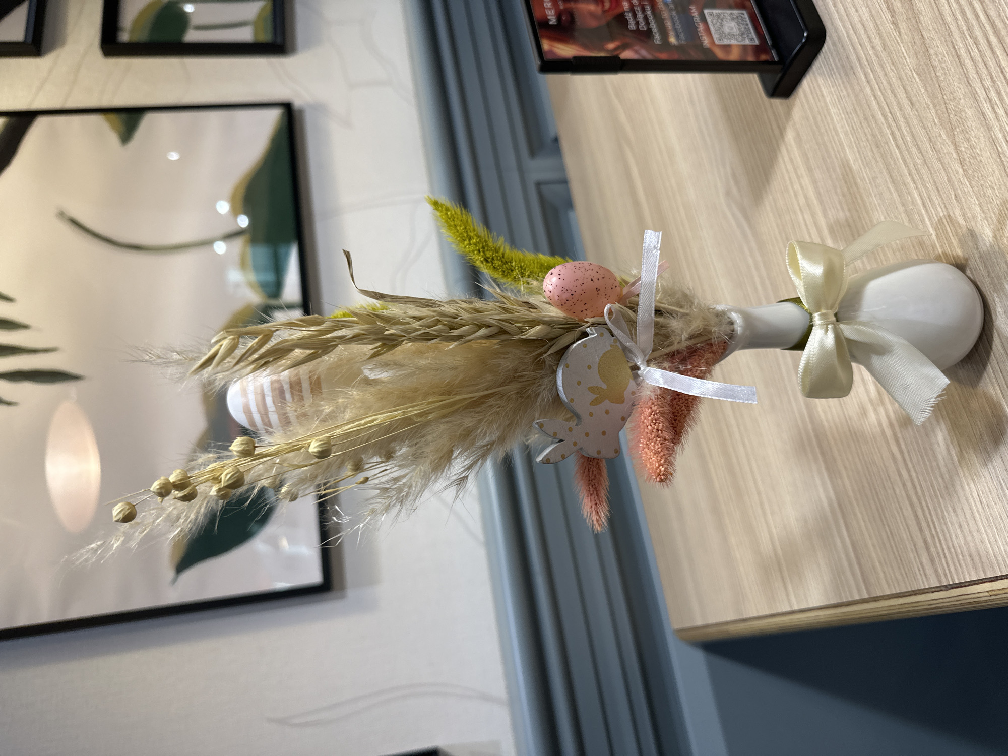 The image shows an Easter decoration on a wooden surface. The decoration consists of dried grasses, a speckled pink egg, a wooden bunny ornament, and ribbons tied around a white, egg-shaped base. In the background, there is artwork on a wall and a textured wall panel. The scene is likely indoors, possibly in a cafe or restaurant. There is no indication of people, actions, or interactions. The location is Wrocław, Poland, according to metadata, but this is not visibly evident in the image itself.
