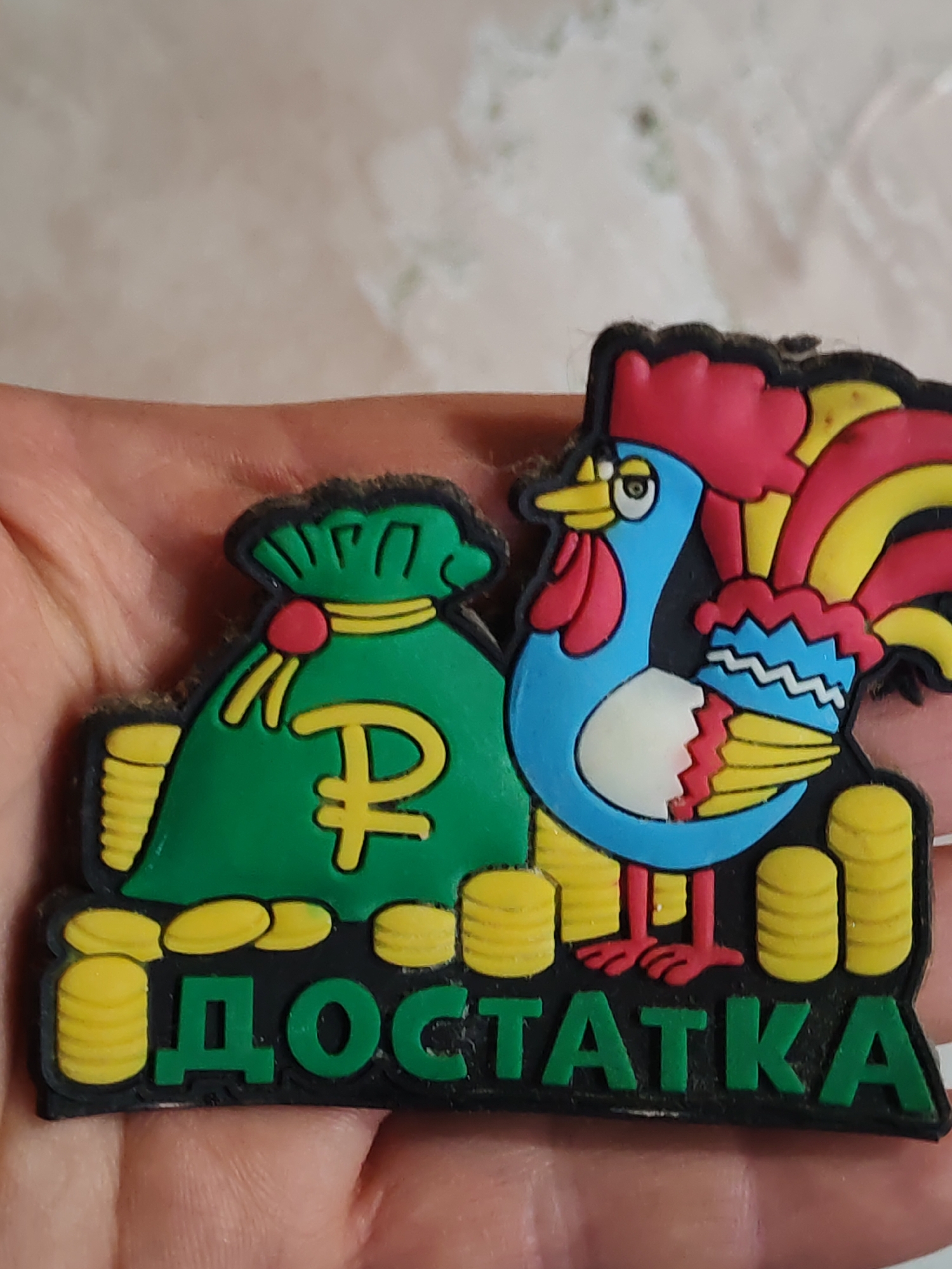 The image shows a rubberized sticker held in a person's hand. The sticker features a green money bag with a yellow ruble symbol, stacks of yellow coins, a blue and red rooster, and the Russian word "ДОСТАТКА" (Dostatka) written in green letters. The word "Dostatka" translates to "abundance" or "prosperity" in English.