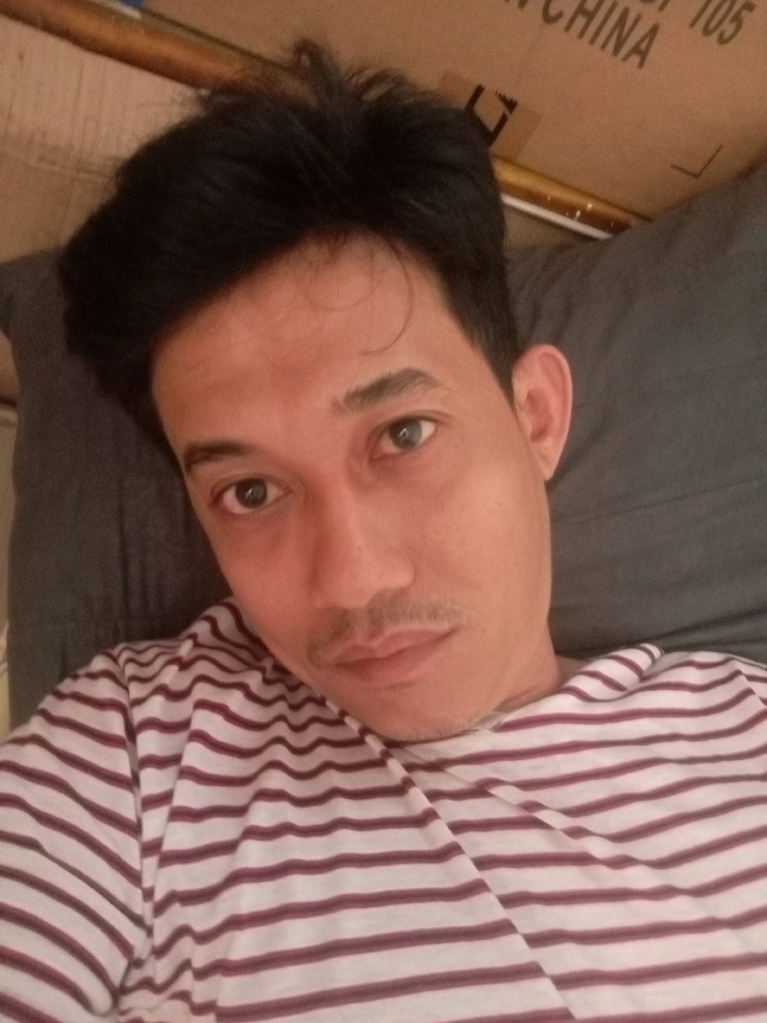 The image is a close-up, overhead shot of a man lying down, looking at the camera. He has dark hair, brown eyes, and a light mustache. He is wearing a white and maroon striped t-shirt. He appears to be in a bedroom, with a pillow behind his head and a box with text that reads "CHINA 105" in the background. The lighting suggests it is daytime. No specific location in Batam, Indonesia, can be determined from the image.