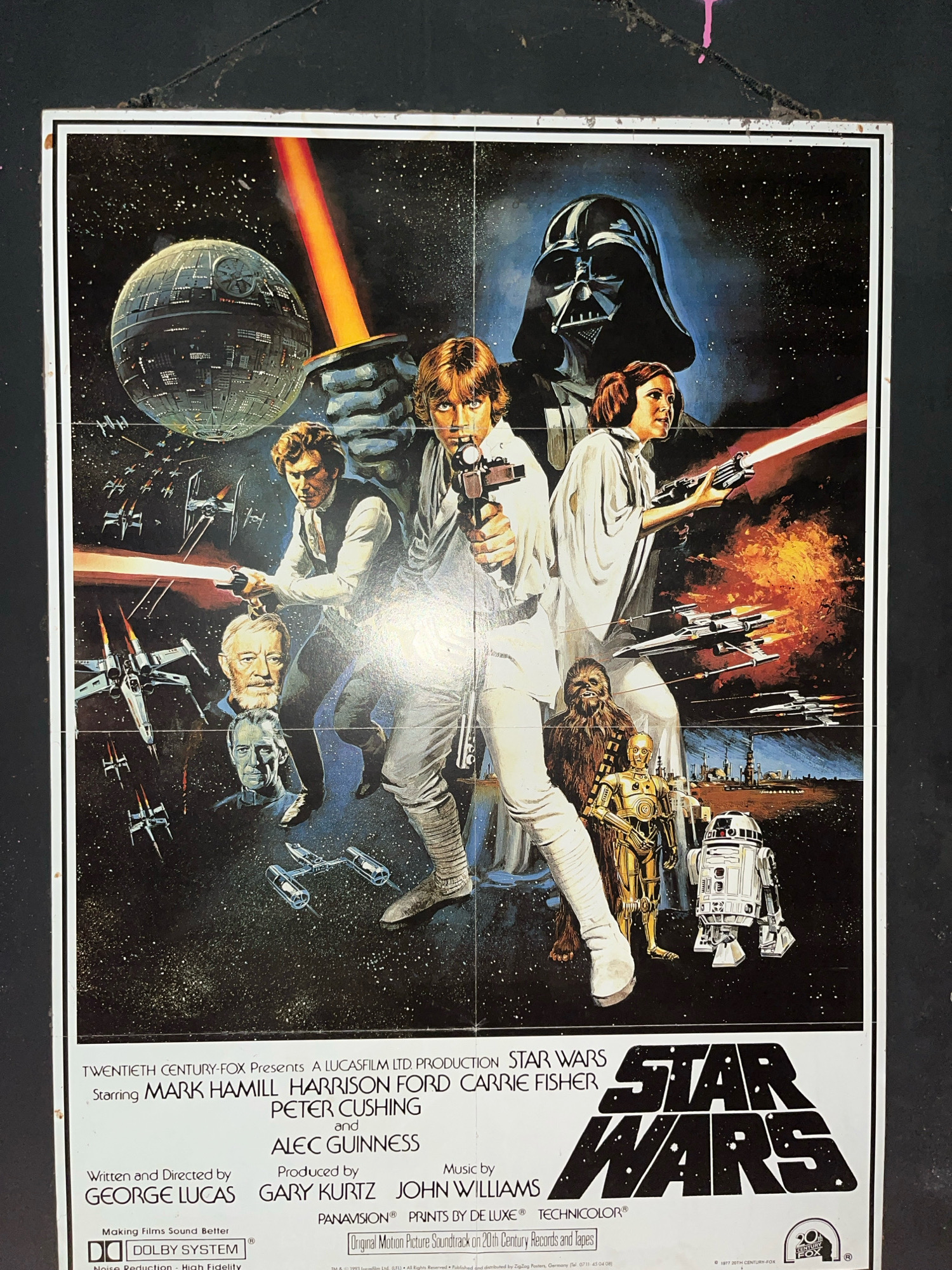 The image is a close-up, eye-level, vertical shot of a vintage "Star Wars" movie poster, likely displayed on a wall. The poster depicts a cosmic battle scene set against a starry black background. Dominating the foreground are the main characters: Luke Skywalker wielding a lightsaber, Princess Leia with a blaster, Han Solo aiming a blaster, and Darth Vader in the upper center. Also visible are the droids C-3PO and R2-D2, the Wookiee Chewbacca, and the characters Obi-Wan Kenobi and Grand Moff Tarkin in portrait form. Numerous starships, including X-wings and TIE fighters, are engaged in combat, with the Death Star visible in the upper left quadrant. The poster has a weathered appearance, with some visible creases and what appears to be pink paint or marker smudges in the upper corners, suggesting it is an older, possibly worn, item. The bottom portion of the poster displays text detailing the cast and crew, production company logos (Twentieth Century Fox, Lucasfilm Ltd., Panavision, Dolby System), and the iconic "Star Wars" logo. The poster is framed by a white border, and the background surrounding the poster is dark, with a rough texture. There is no discernible location in the city of Surakarta, Indonesia.