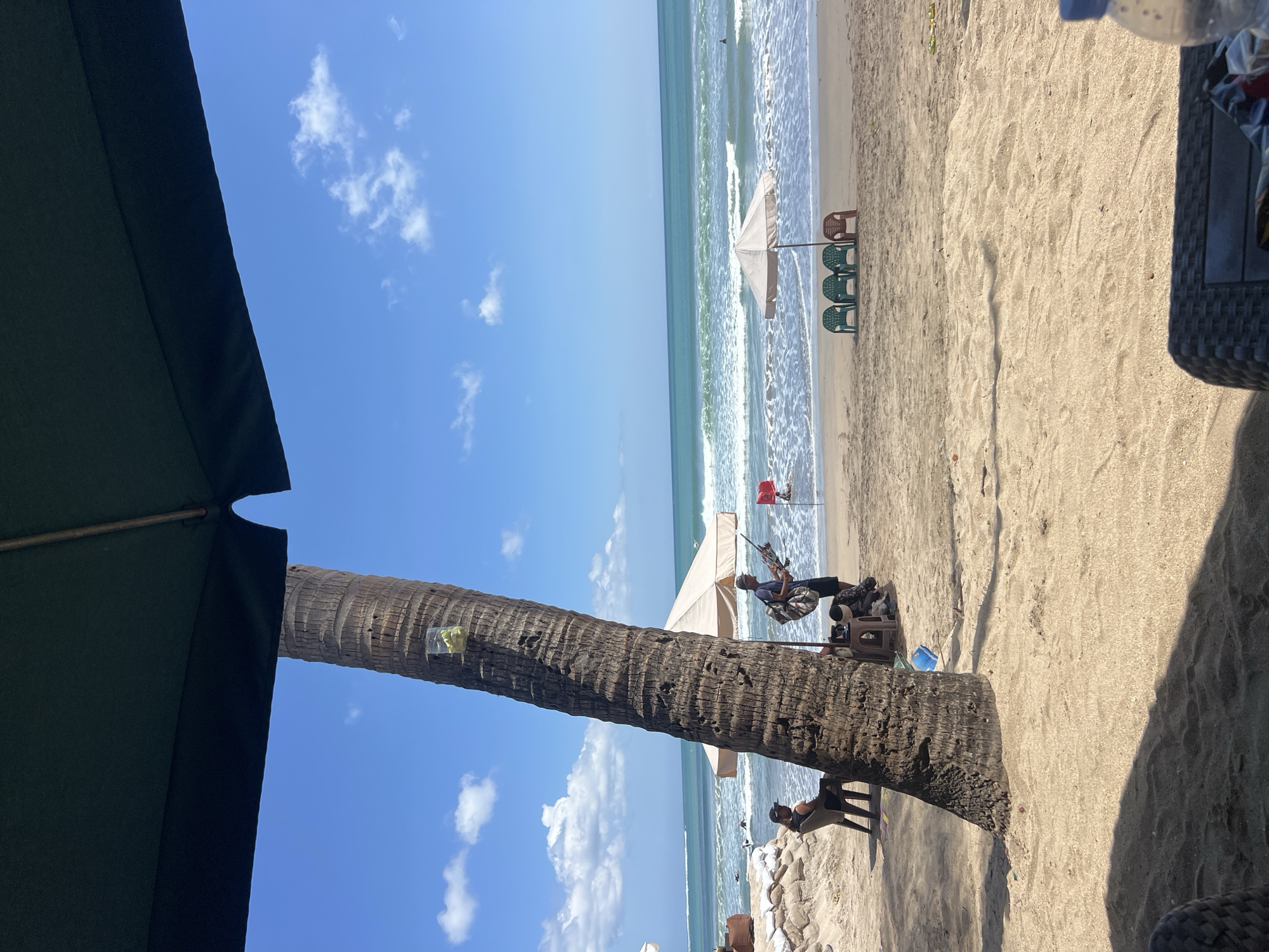 This is a wide-angle photograph taken on a sunny day at Kuta Beach, Indonesia. The foreground is dominated by sand and the shadow of a large umbrella overhead. A palm tree trunk is prominent on the left side of the frame, with a clear plastic bag containing some items hanging from it.
In the midground, the sandy beach slopes down towards the ocean. Several people are present. On the left, a person sits on a chair near a sandbag embankment. Closer to the water, another person stands with what appear to be fishing rods. There is a small beige umbrella providing shade for a table with a blue plastic bag and another object underneath. Further out on the beach, three green plastic chairs and one brown plastic chair are arranged in front of another beige umbrella, near the water's edge.
The ocean is turquoise with white-capped waves breaking on the shore. In the distance, a few surfers or swimmers are visible in the water. The sky is a vibrant blue with scattered white clouds. The scene depicts a typical day at a popular tourist beach.