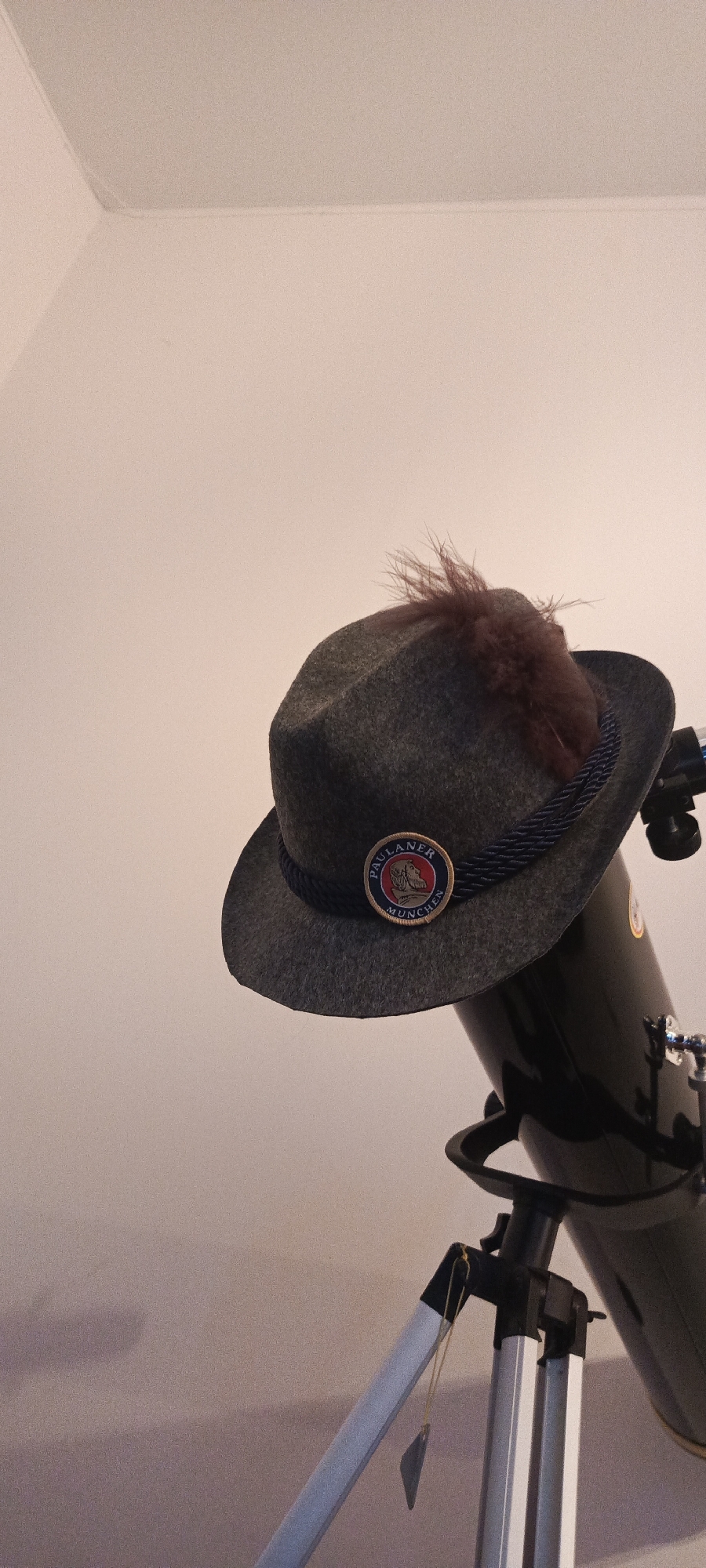 A dark grey felt hat with a dark blue braided band and a brown feather adorns the top of a black telescope, which is mounted on a silver tripod. A circular patch with red, blue, and white detailing is affixed to the front of the hat. The background is a plain, light-colored wall, possibly in a room. The overall scene suggests a still-life arrangement, with the hat serving as an unusual accessory for the telescope. The image does not provide any specific details that would allow for identification of its location in Rustavi, Georgia.