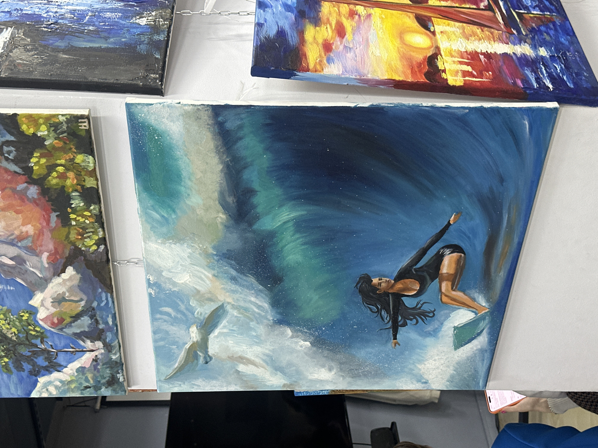An eye-level shot captures multiple paintings displayed on what appears to be a white surface, likely at an art exhibition or market in Sochi, Russia.
The central and most prominent artwork is a dynamic painting of a woman surfing a large wave. She has long dark hair, wears a black, long-sleeved swimsuit, and is depicted in an active, balanced pose on her surfboard, riding the crest of a breaking wave. The ocean water is painted in various shades of deep blue and turquoise, with white foam and spray, and subtle speckles suggesting water droplets or light reflection. Two white seagulls are visible flying near the wave's white crest on the left side of the painting.
Surrounding this central piece are parts of other artworks. To the top left, a painting features dark blues and blacks with white highlights, possibly depicting a stormy sea or an abstract landscape. Above and to the right, a vibrant painting shows warm yellows, oranges, and reds against a darker background of blues and purples, reminiscent of city lights reflecting on water or a sunset scene. To the bottom left, a landscape painting is partially visible, showing green foliage, blue water, and light-colored rock formations. Faint letters, possibly "TH," are visible on the top edge of this landscape painting.
The canvases are either propped up or hanging, with a small metal chain visible between two of the upper paintings. The overall lighting is bright and even. In the bottom right corner, a person's hand holding a red-cased smartphone is partially visible, indicating presence at the display.