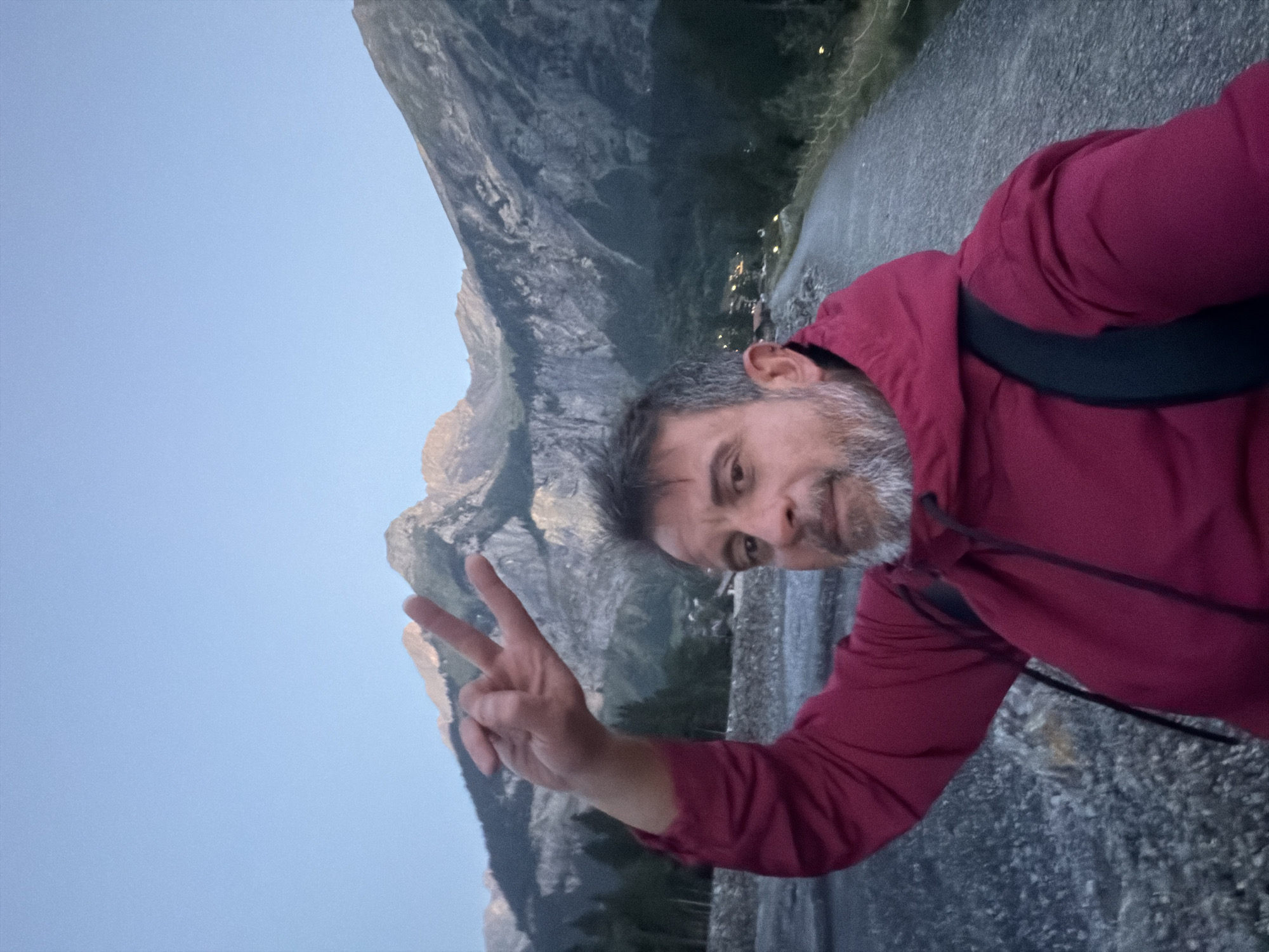 In Kandersteg, Switzerland, at dusk, a middle-aged man with graying hair and beard takes a selfie. He is wearing a dark red hooded jacket and a black backpack strap is visible. He is holding up his right hand in a peace sign. The man is standing on a gravel path with a rocky embankment to his left. In the background, large, rugged mountains dominate the scene. The sky is a clear, pale blue, transitioning to a deeper blue at the top, indicative of twilight. A few scattered lights can be seen in the valley between the mountains, suggesting a small settlement. Trees line the lower slopes of the mountains on the left side of the frame.