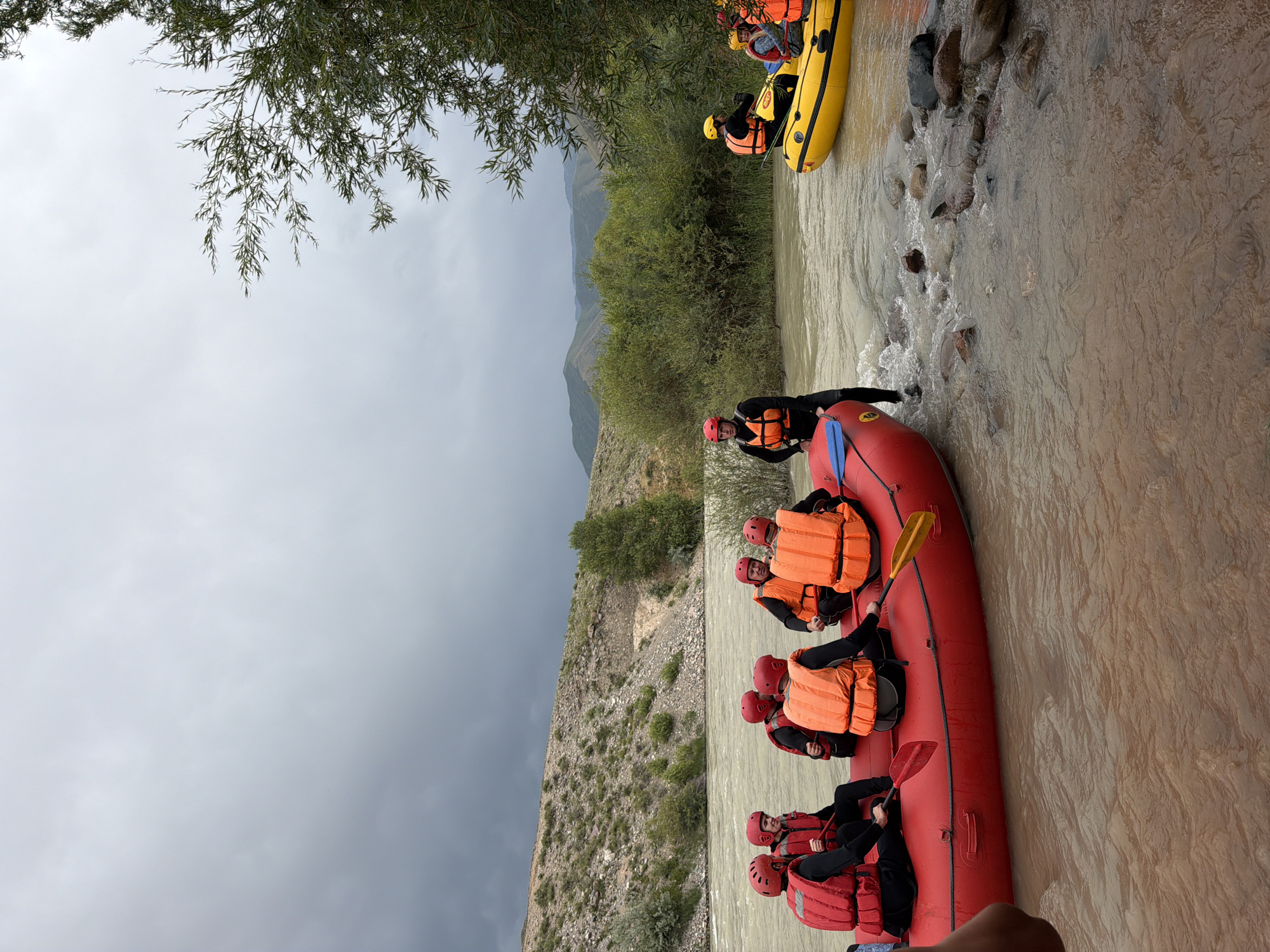 In Sasyk-Bulak, Kyrgyzstan, a group of people are preparing for a rafting excursion on a cloudy day. Two inflatable rafts are in the water near the shore. A large red raft, with six individuals wearing red and orange life vests and helmets, is partially submerged in the shallow water. Some individuals are holding paddles. To the right, a smaller yellow raft, with two individuals also wearing life vests and helmets, is further out in the river. The river water is murky brown, with visible rocks in the foreground. The surrounding environment consists of a rocky, vegetated hillside and lush green trees and bushes. In the distance, rolling hills under a gray, overcast sky complete the scene.