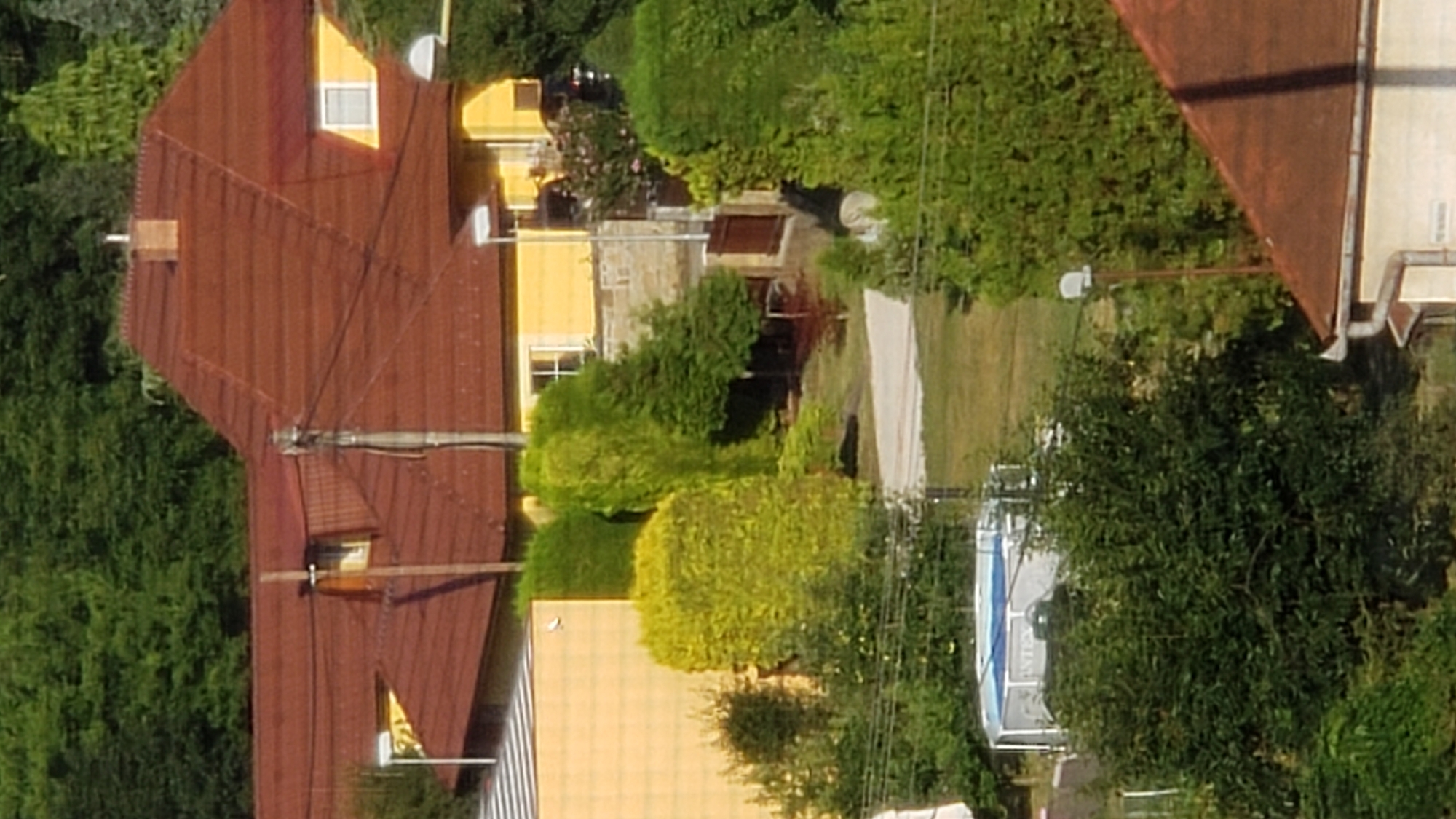 This image depicts a residential area with a prominent yellow house featuring a dark red, tiled roof. The house has multiple windows, one of which is visible on the upper floor. A brick chimney is present on the roof. The foreground and surrounding area are filled with lush green trees and bushes, some of which are manicured into rounded shapes. A light-colored pathway or driveway winds through the greenery. In the lower left quadrant, a round, above-ground swimming pool with a blue interior and a silver-colored frame is visible. Several utility poles with wires are scattered throughout the scene. The overall impression is of a suburban or semi-rural setting. The provided crops suggest the location may be within Prešov, Slovakia, but this cannot be definitively confirmed from the image alone.