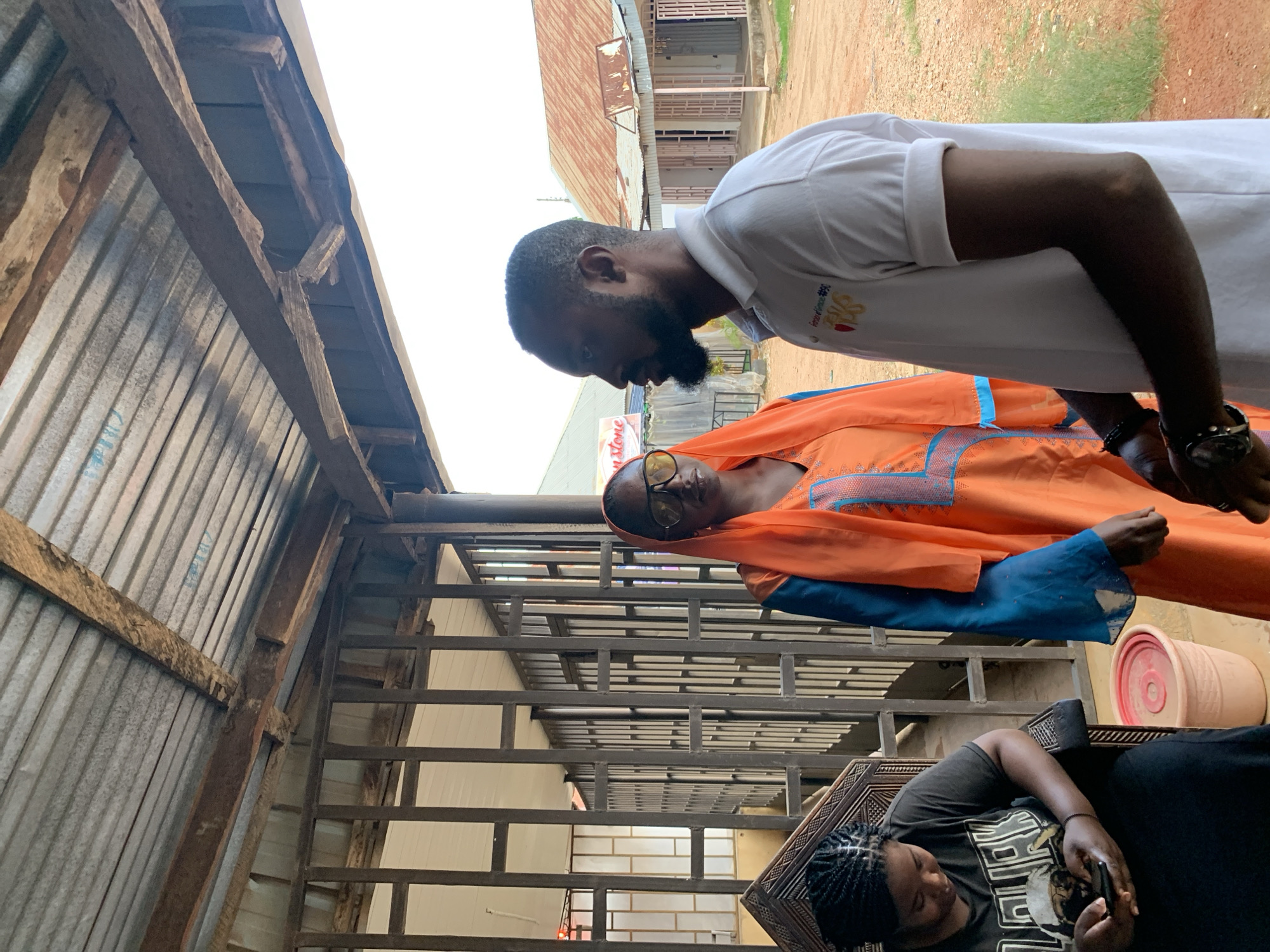 In Jos, Nigeria, three people are gathered under a corrugated metal roof supported by wooden beams. On the left, a woman with braided hair sits in a chair, looking at her phone. To her right, a woman in an orange hijab and a blue-sleeved garment stands, wearing large sunglasses. Facing the woman in the hijab is a man wearing a white polo shirt. The background reveals buildings with corrugated metal roofs and barred windows, suggesting a commercial or institutional setting. The ground is covered in reddish dirt and patches of green grass.