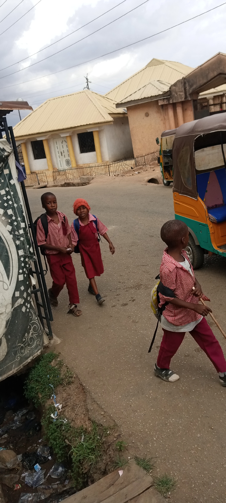 Three children in school uniforms walk on a paved road in Jos, Nigeria. Two children are walking together, one in a red dress and one in a red shirt and pants. The third child walks alone with a stick. An auto rickshaw is parked in the background. Buildings with beige roofs are visible. A gutter filled with trash and vegetation runs along the side of the road.