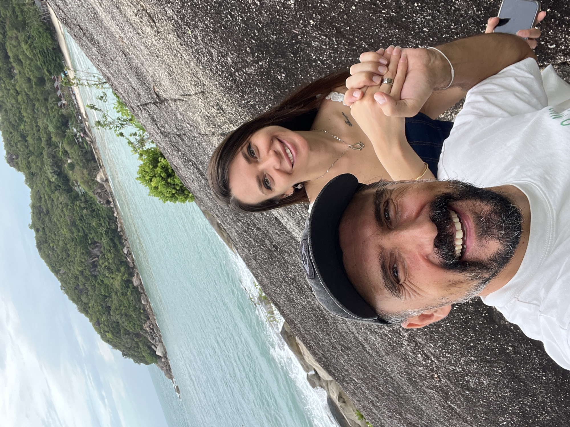 A couple poses for a selfie. The woman has long brown hair and is wearing a navy top. The man wears a baseball cap and a white t-shirt. They are holding hands. In the background is a body of water, a rocky outcropping with lush green vegetation, and a beach with buildings. The photo was taken on a large rock surface. The location is Baan Chaweng Noi, Thailand.