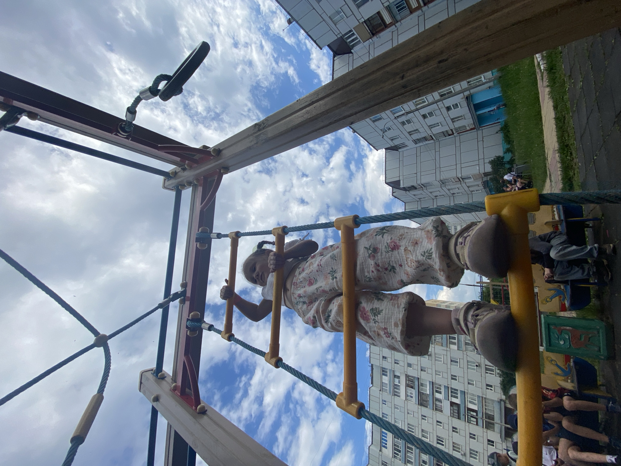 A low-angle shot captures a young girl climbing a rope ladder on a playground. The girl, wearing a white top and floral patterned pants, is partially visible as she ascends the ladder, her hands gripping the yellow rungs and her feet on the ropes. Her hair is tied in pigtails.
The playground equipment is made of metal and wood, with thick ropes forming the ladder. Behind the playground, tall, multi-story apartment buildings are visible, suggesting an urban or suburban residential setting. The sky above is blue with scattered white clouds, indicating daytime, likely fair weather. Some other people are visible sitting on a bench in the background, out of focus.
The scene is likely taking place in Chaikovsky, Russia, given the context. The overall mood appears to be playful and active, focused on the child's activity. There is no visible text in the image.
