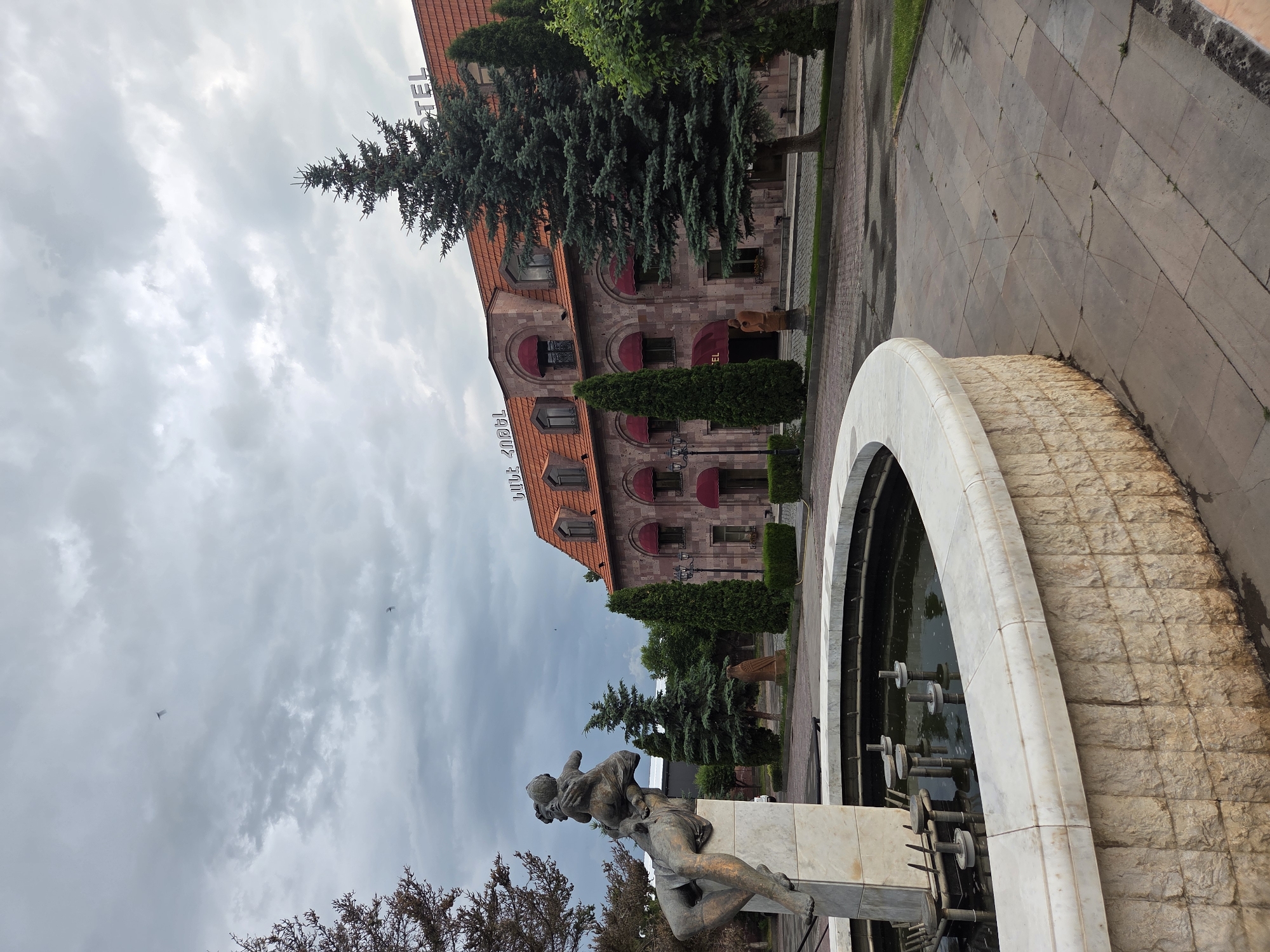 The image shows a monument and a building on a cloudy day. In the foreground, there is a large stone fountain with a sculpture on top. The sculpture depicts a person holding a child. The fountain is circular and made of stone blocks. Behind the fountain, there is a building with a red tiled roof and arched windows. The word "HOTEL" is visible in large letters on the roof of the building. The building is made of stone and has red awnings over the windows. There are several trees and bushes around the building and fountain, including a large evergreen tree in front of the building. The ground around the fountain and building is paved with stone tiles. The sky is cloudy and gray. The location is stated to be in the city of Border Gyumri-Akhuryan, Armenia.