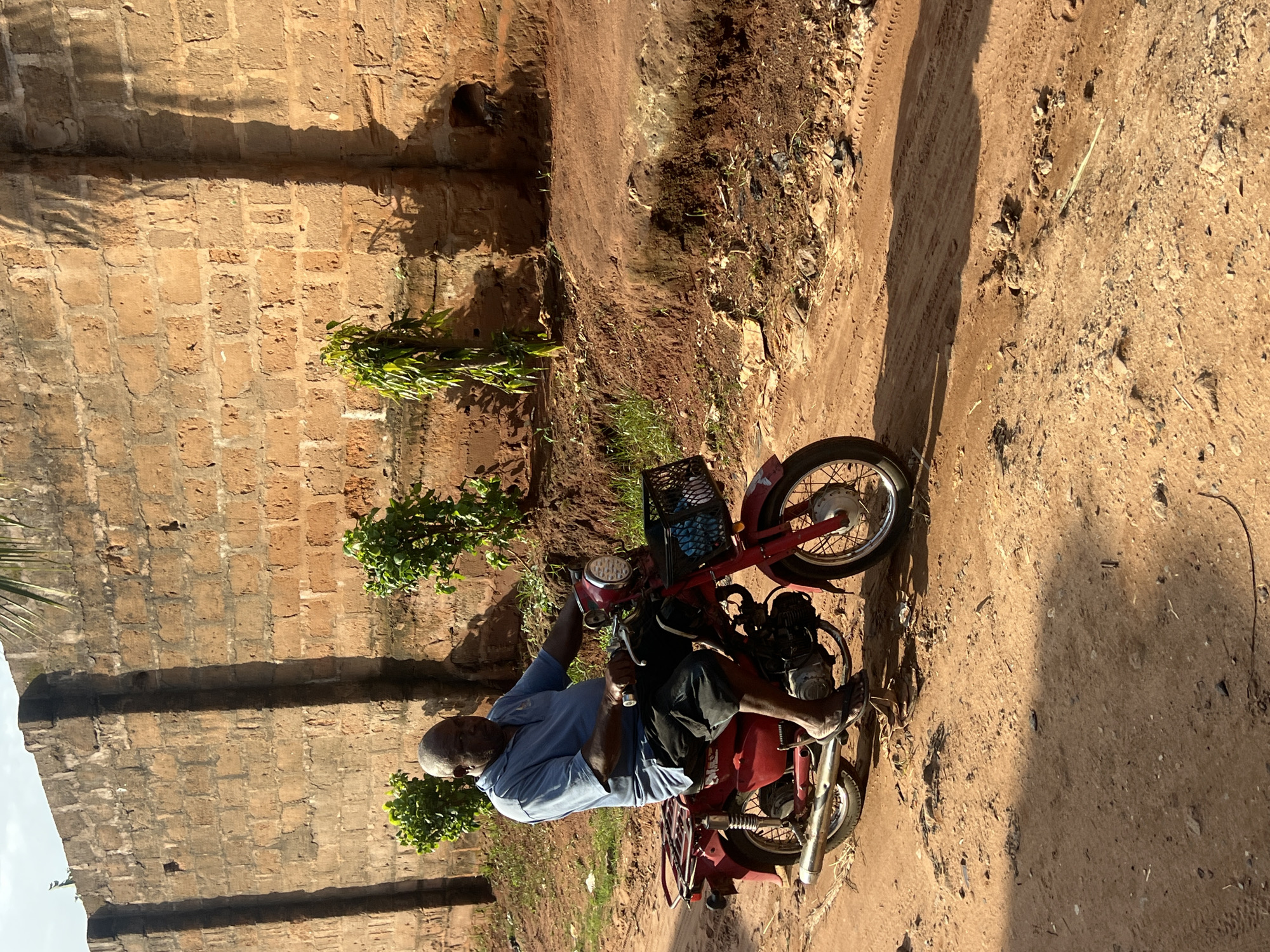 A man sits on a red motorcycle in Nkpor, Nigeria. He is wearing a light blue collared shirt and dark pants, with his left leg extended forward and his right leg bent. The motorcycle has a basket on the front and a rack on the back, and its engine is visible. The man appears to be either preparing to ride or has just stopped.
The scene is outdoors, with a textured brick wall on the left side of the image and a dirt path with tire tracks on the right. Sparse green vegetation grows near the wall and along the path. The lighting suggests it is daytime, with strong sunlight casting shadows. The overall impression is of an everyday scene in a rural or semi-rural environment. There is no visible text in the image.