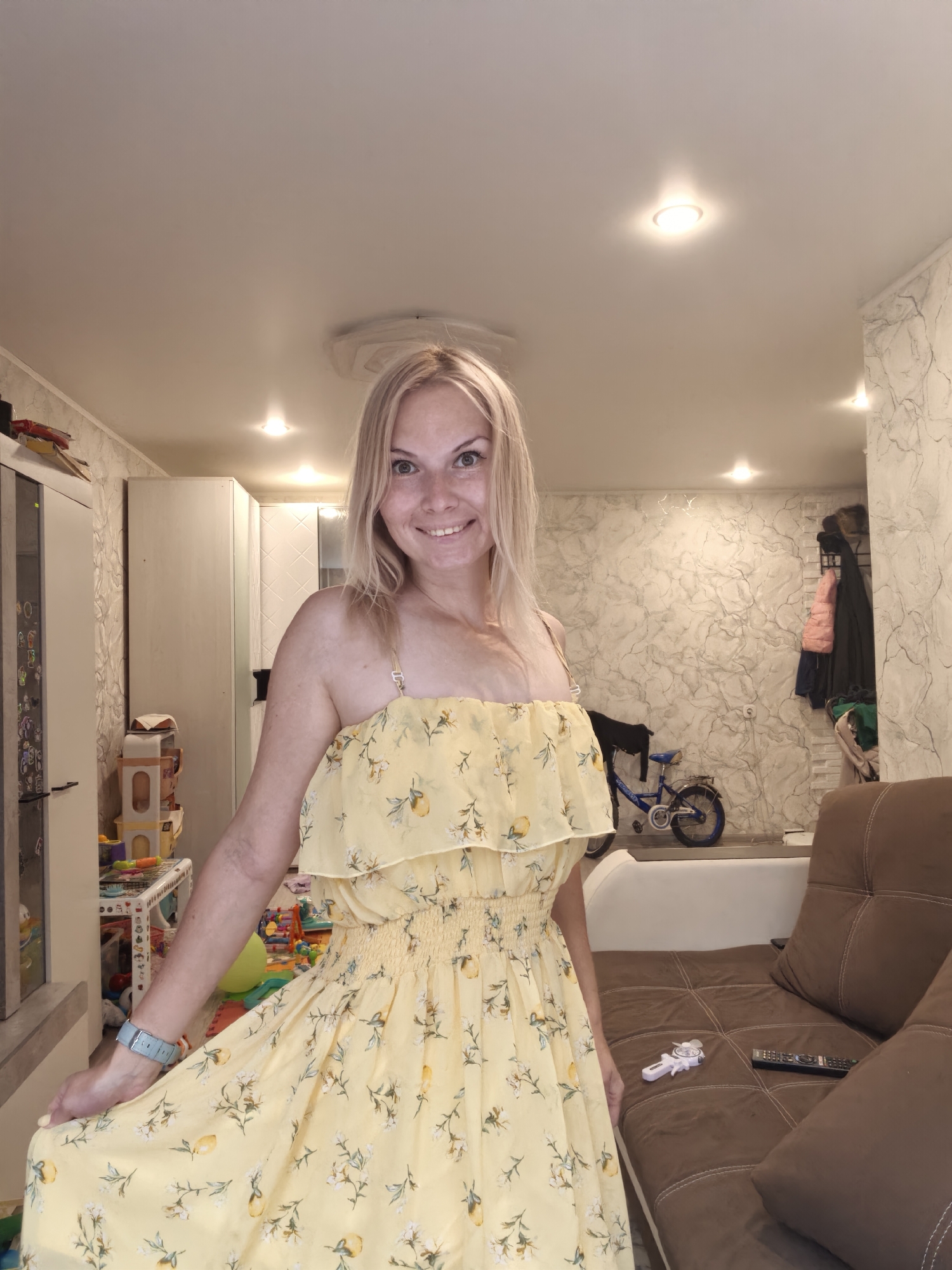 A blonde woman wearing a yellow floral dress stands in a living room. She is smiling at the camera. The room contains a sofa, a coffee table with a remote control and a toy, a bicycle, and shelves with various items. The walls are decorated with textured wallpaper, and the ceiling has recessed lighting. The woman is on the left side of the frame, and the sofa is on the right. There is a window on the left side of the room, and a doorway in the background. The overall impression is that of a casual indoor setting.